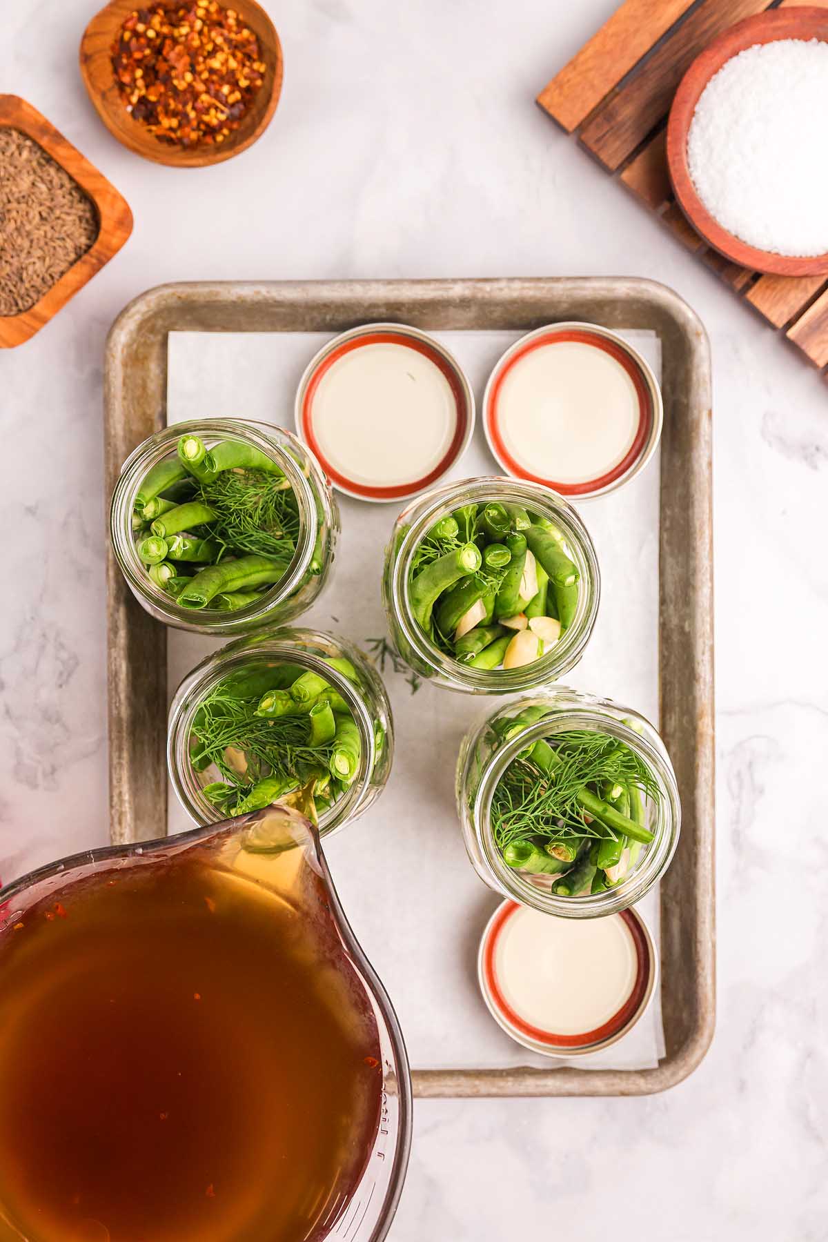 Brine being poured into jars with pickled green beans, herbs, and garlic, surrounded by jar lids and spices.