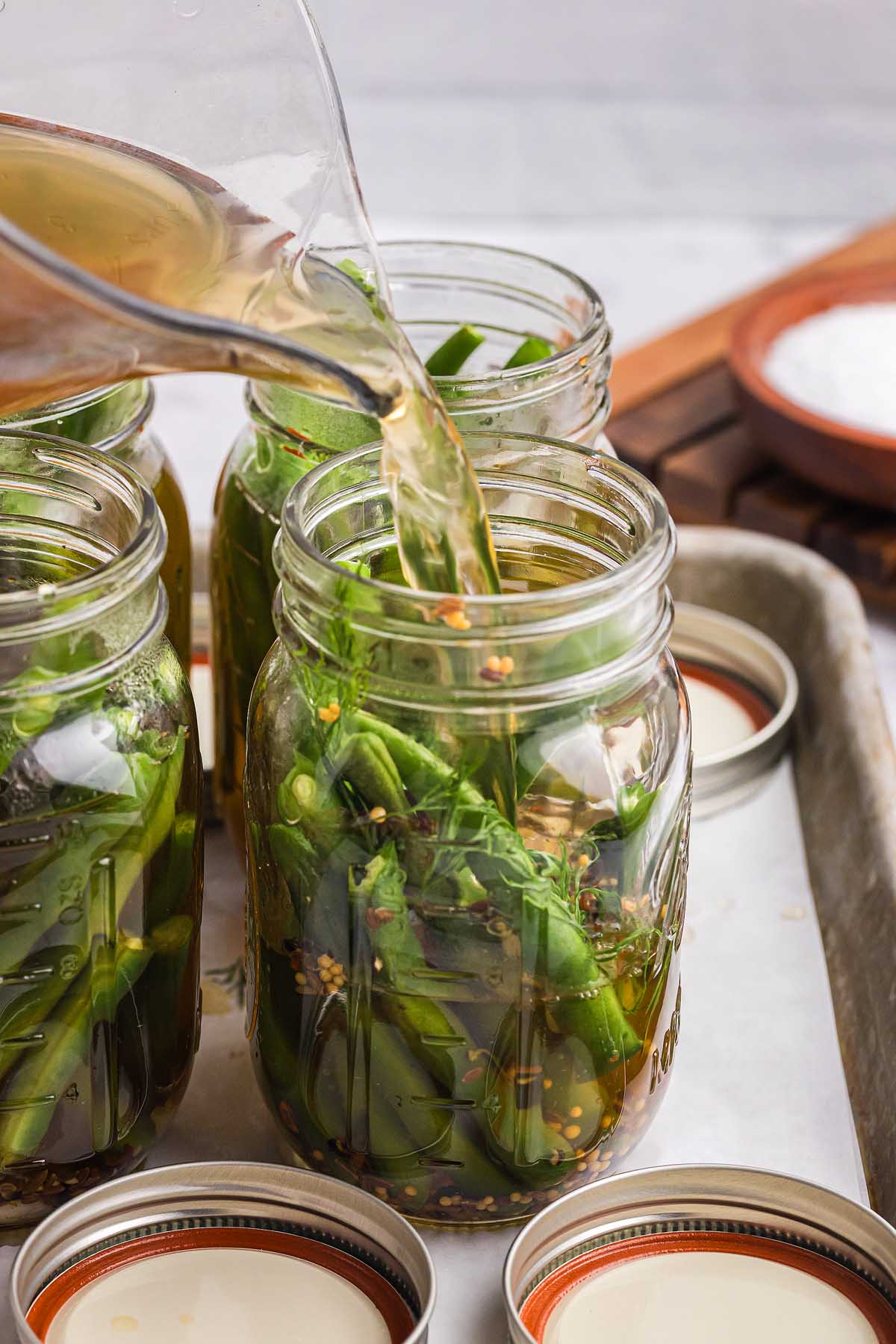 Brine is being poured into jars filled with fresh green beans and spices, creating delicious pickled green beans ready for preserving.