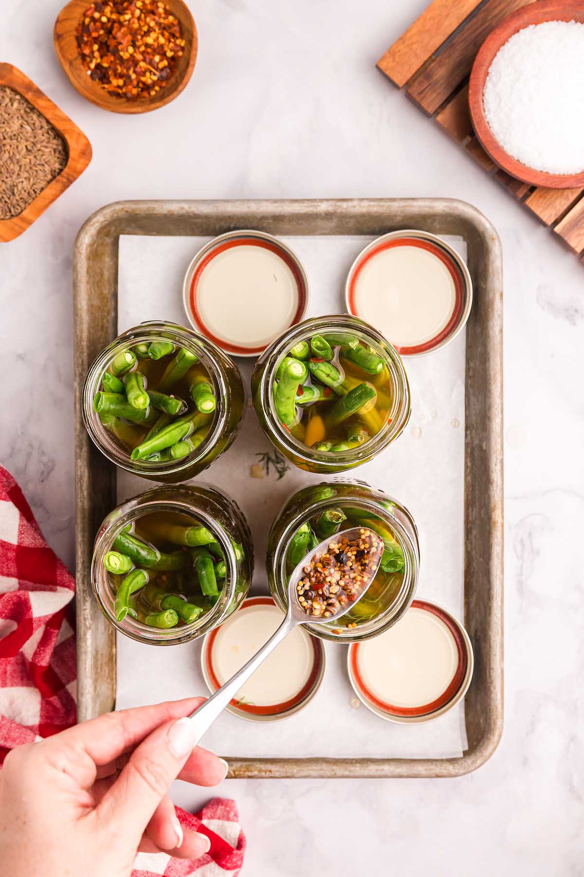 A hand adds spices to jars of pickled green beans in brine on a tray, with salt and seasonings nearby.