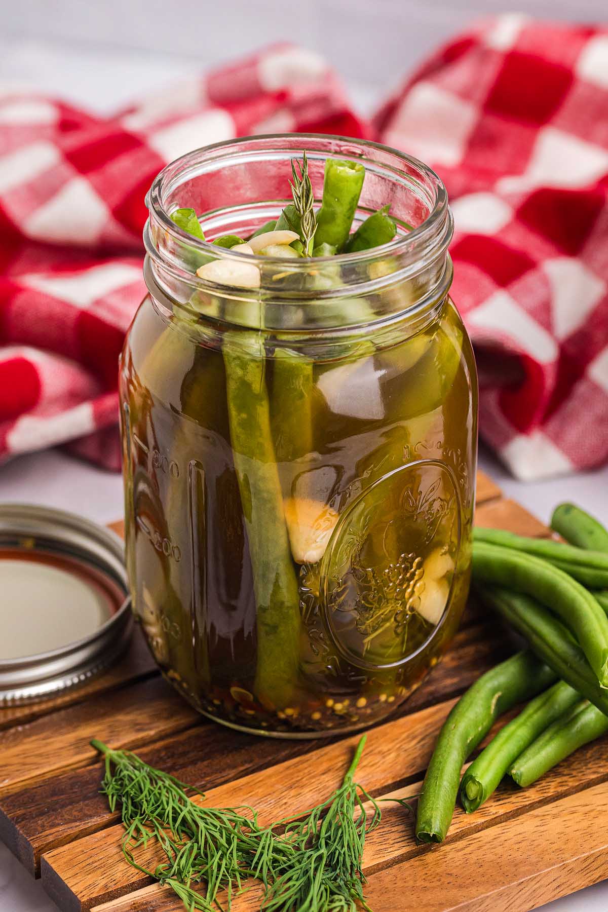 A glass jar of pickled green beans with garlic and dill sits on a wooden board, surrounded by fresh green beans and dill, highlighting the crisp texture and tangy flavor of these homemade pickled green beans.