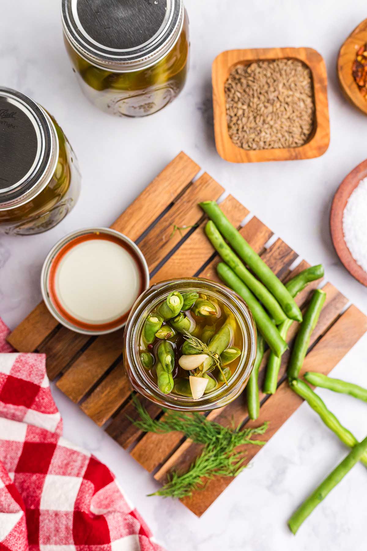 Open jar of pickled green beans on a wooden board, surrounded by fresh beans, spices, and a red checkered cloth—perfect for showcasing homemade pickled green beans.