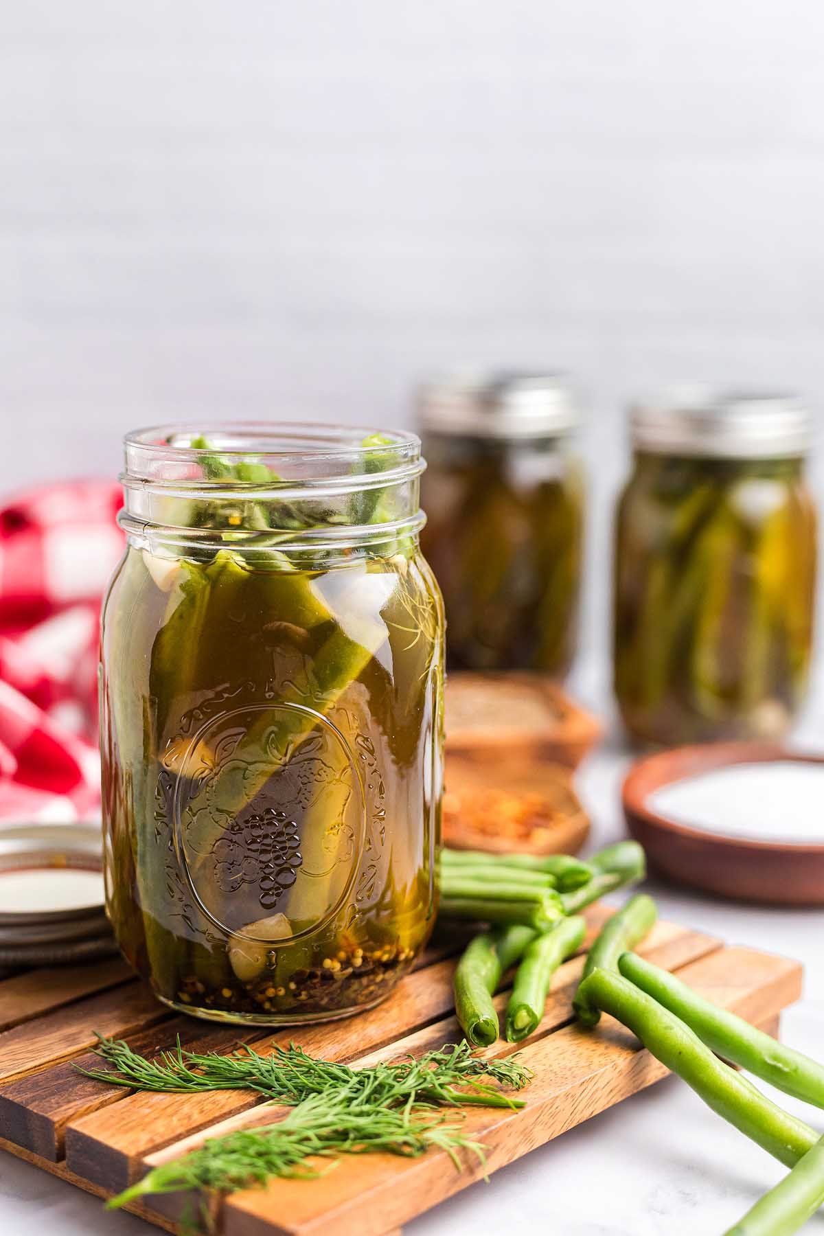 A jar of pickled green beans sits on a wooden board, surrounded by fresh beans, dill, jars, and spices in the background.