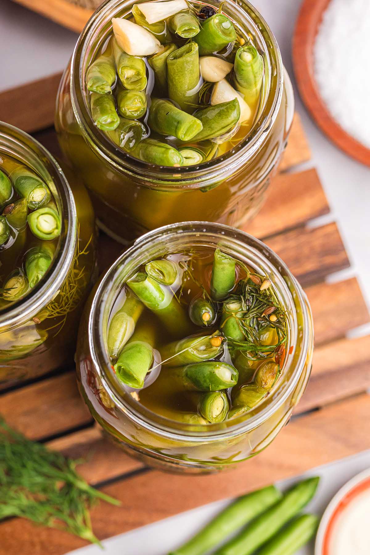 Jars filled with pickled green beans, fresh herbs, and brine rest on a wooden surface.