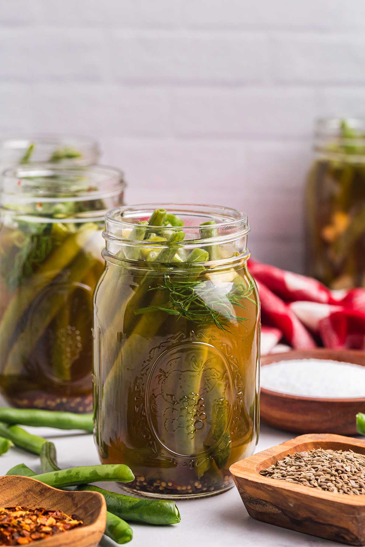 A mason jar filled with pickled asparagus spears and pickled green beans, surrounded by spices and more jars in the background.
