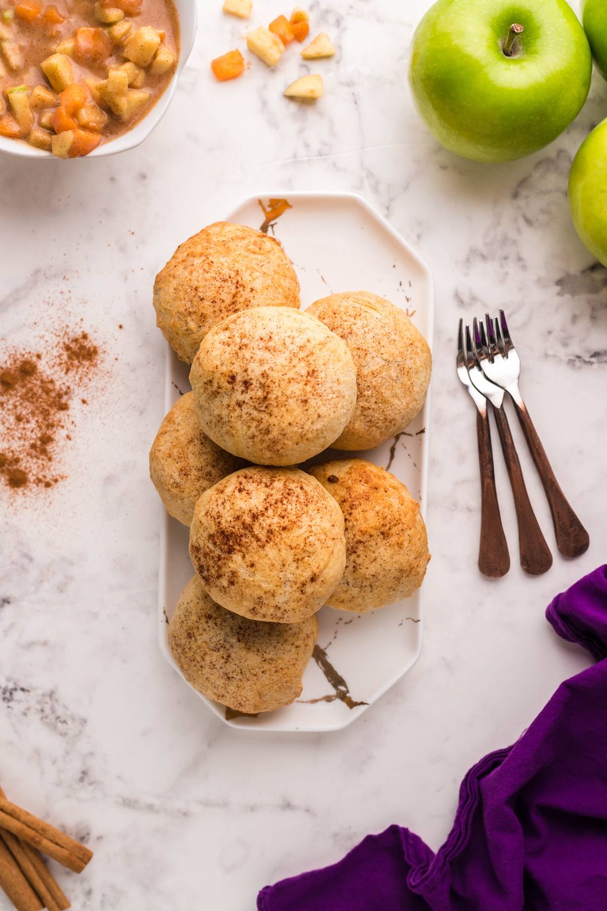 Round Air Fryer Apricot Apple Dumplings dusted with cinnamon on a white platter, surrounded by apples and forks on a marble surface—an easy way to see how to make dumplings with puff pastry for a delicious treat.