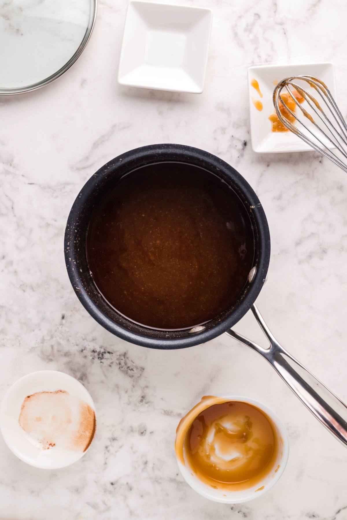 A saucepan of caramel sauce on a marble counter, with bowls, a whisk, and caramel remnants around it—perfect for drizzling over autumn air fryer desserts like Air Fryer Apricot Apple Dumplings.