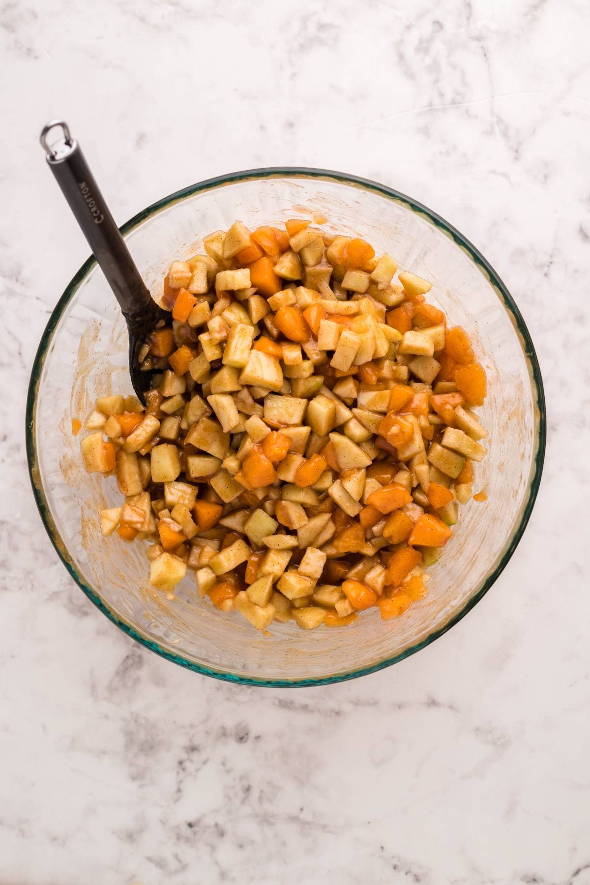 A glass bowl with diced apples and apricots being mixed with a spoon on a marble countertop—perfect for creating autumn air fryer dessert or preparing filling for puff pastry dumplings.