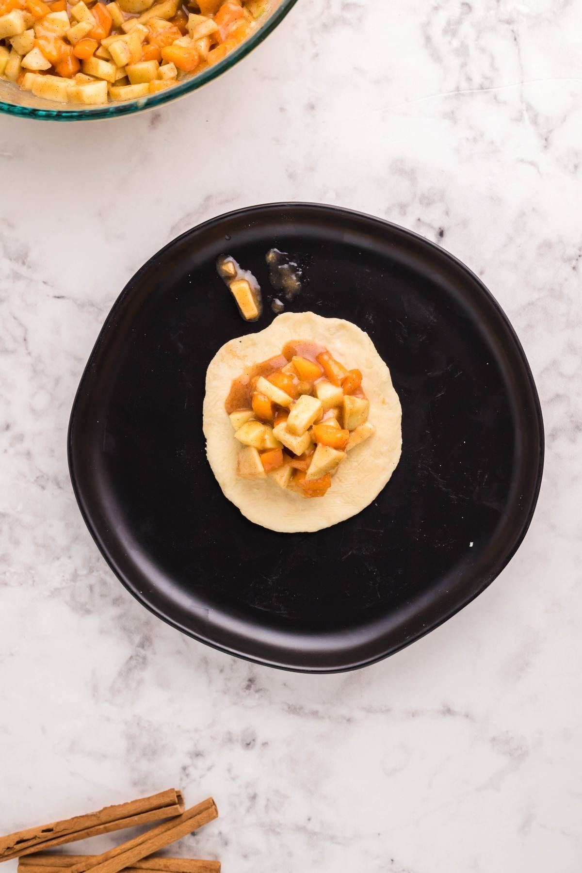 Unbaked dough circle on a black plate topped with diced apple filling, hinting at delicious Air Fryer Apricot Apple Dumplings, with cinnamon sticks nearby for extra flavor.
