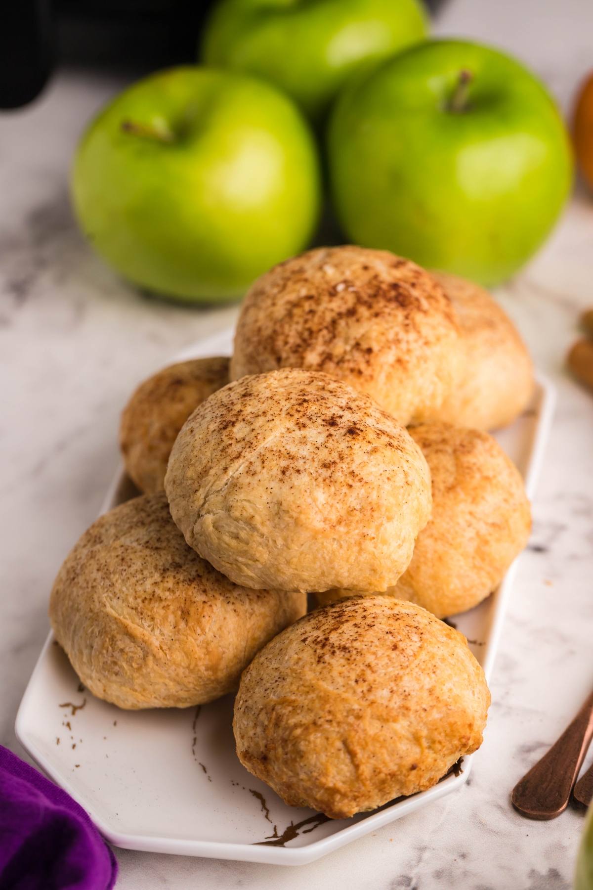 A plate of cinnamon-dusted baked rolls with green apples in the background, reminiscent of Air Fryer Apricot Apple Dumplings—perfect if you want to learn how to make dumplings with puff pastry at home.