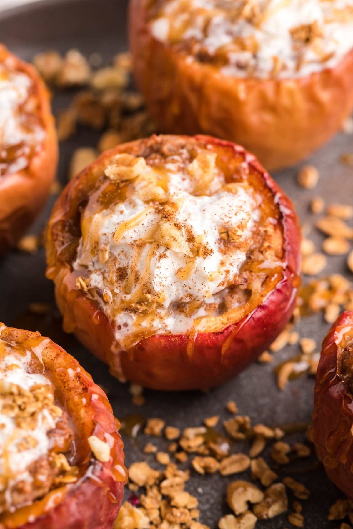 Baked apples stuffed with oats and topped with cream, sprinkled with cinnamon and granola, made with maple syrup for extra sweetness on a dark surface.