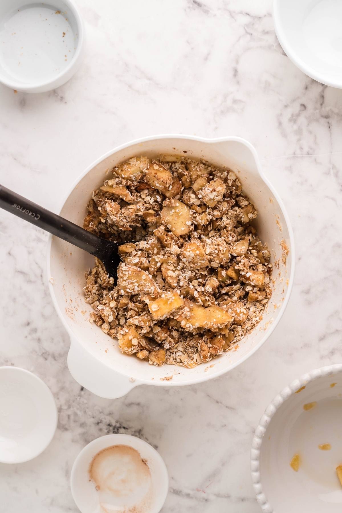 A bowl of apple crisp mixture with oats is being stirred by a black spoon on a marble countertop—perfect for making an air fryer apple dessert or to bake apples in the air fryer for a quick, delicious treat.