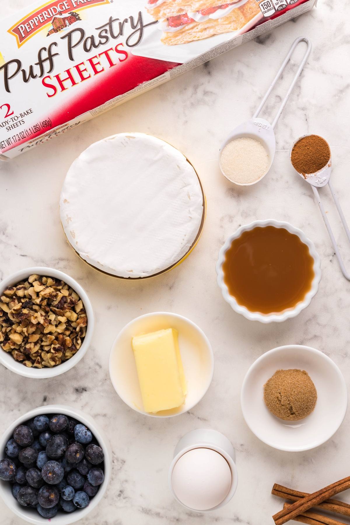 Ingredients on a marble counter: puff pastry, brie cheese, butter, egg, blueberries, nuts, sugars, spices, and syrup—everything you need for a delicious blueberry brie appetizer or Brie and puff pastry baked in an air fryer.