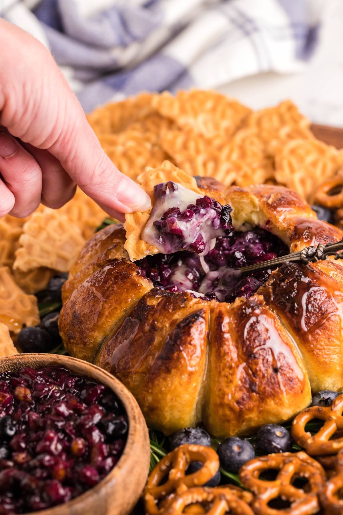 A hand dips a cracker into an Air Fryer Baked Brie with Blueberries, surrounded by pretzels and crackers for a delicious blueberry brie appetizer.