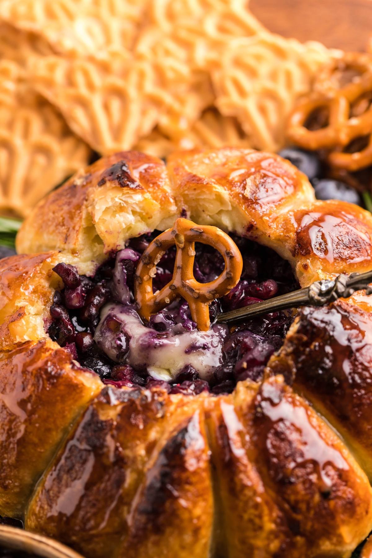 Golden baked pastry filled with melted cheese and berries, topped with a pretzel, served with crackers—experience this Air Fryer Baked Brie with Blueberries for a modern twist on classic flavors.