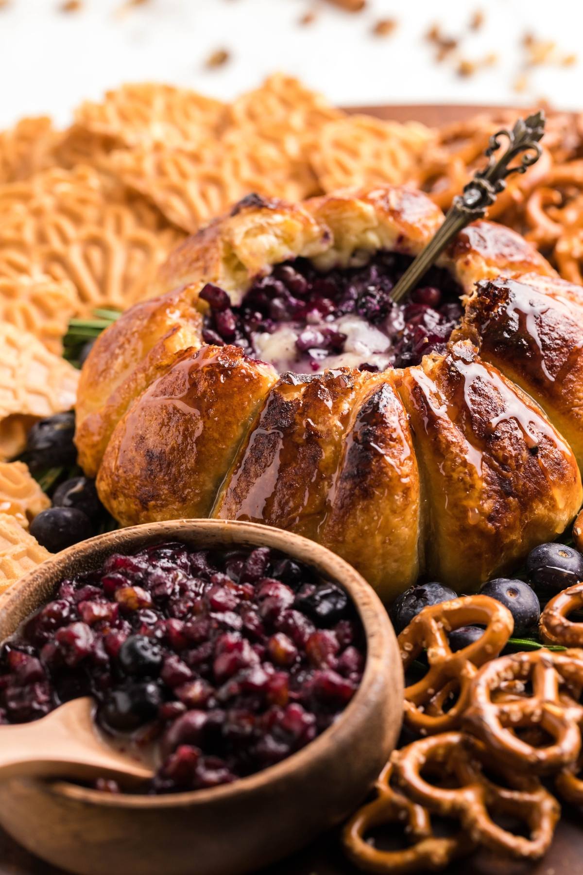 Baked brie wrapped in pastry with berries inside, surrounded by pretzels and crackers on a platter, makes a perfect blueberry brie appetizer.
