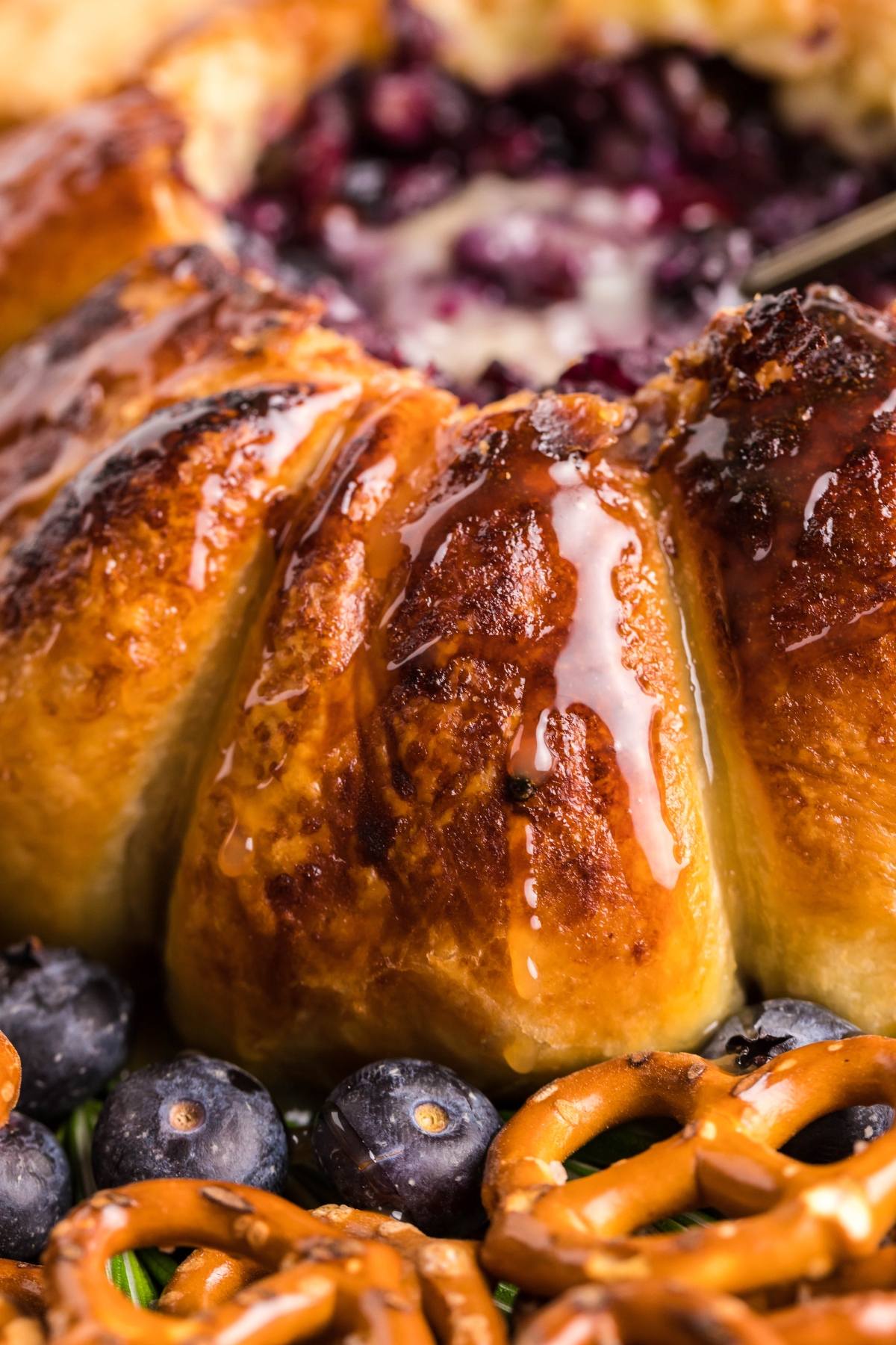 Golden baked pastry with fruit filling, blueberries, and pretzels in the foreground, drizzled with glaze—a delightful blueberry brie appetizer made with Brie and puff pastry baked in an air fryer.