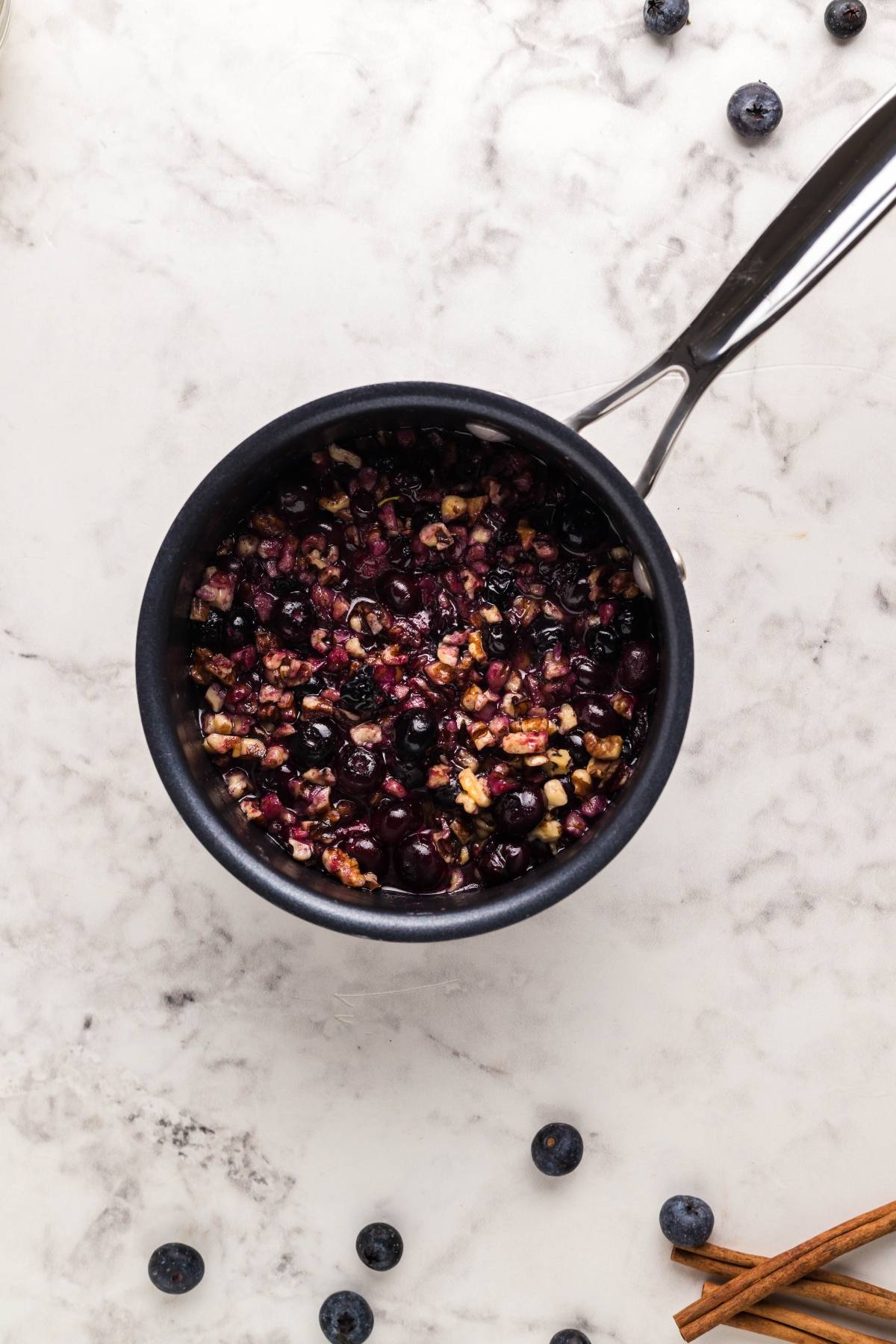 Small saucepan with a blueberry and nut mixture for a delicious blueberry brie appetizer, shown on a marble countertop with scattered blueberries and cinnamon sticks.