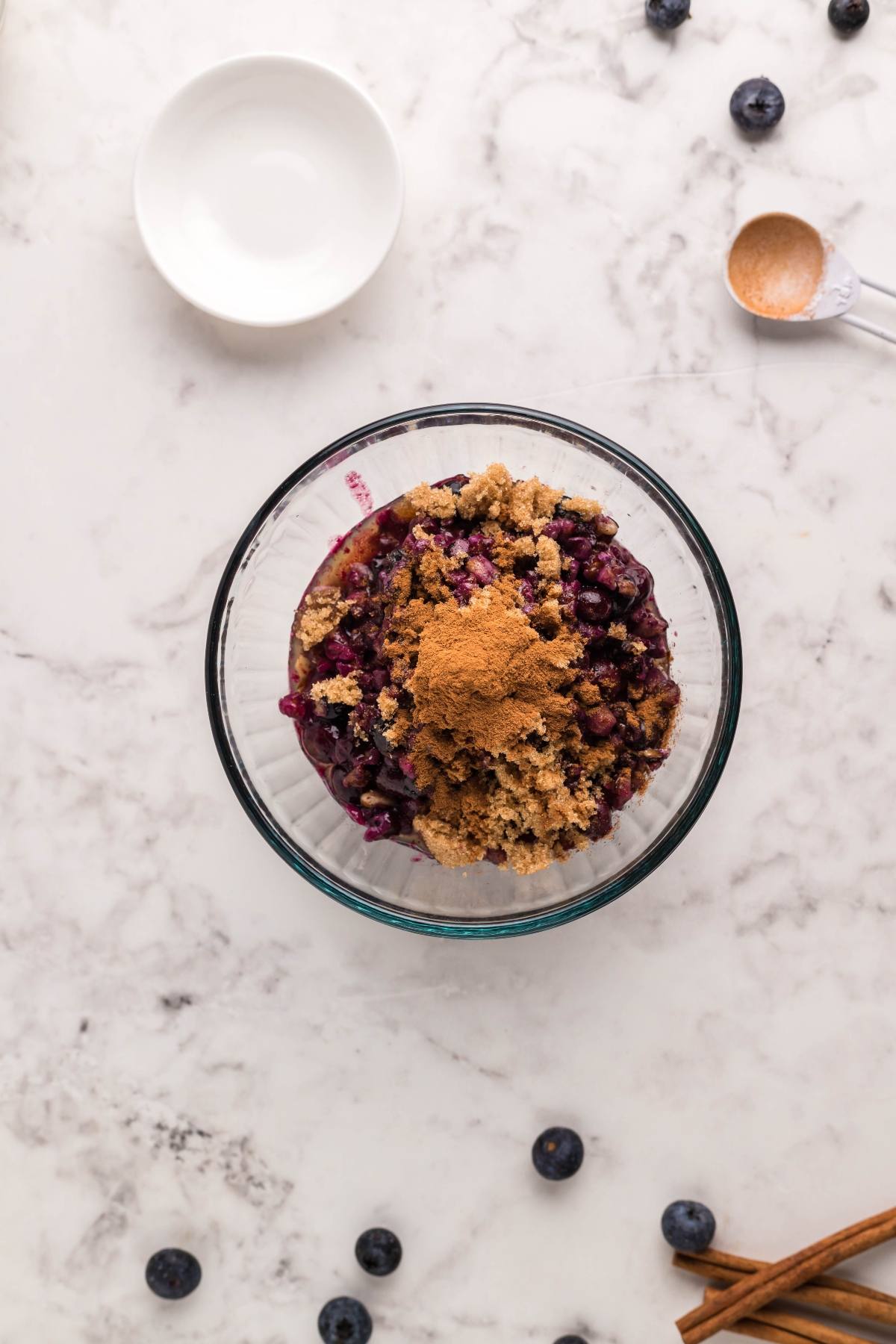 Glass bowl with blueberries, brown sugar, and cinnamon on a marble surface—perfect for topping an Air Fryer Baked Brie with Blueberries or adding flair to your next blueberry brie appetizer. Scattered berries and cinnamon sticks complete the scene.