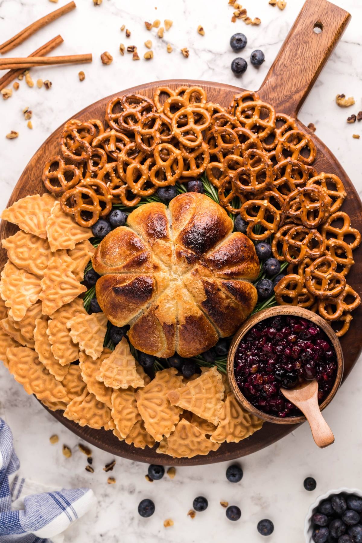 A round serving board with pretzels, waffle cookies, blueberries, a pumpkin-shaped bread, berry jam, and an Air Fryer Baked Brie with Blueberries for a delightful blueberry brie appetizer.
