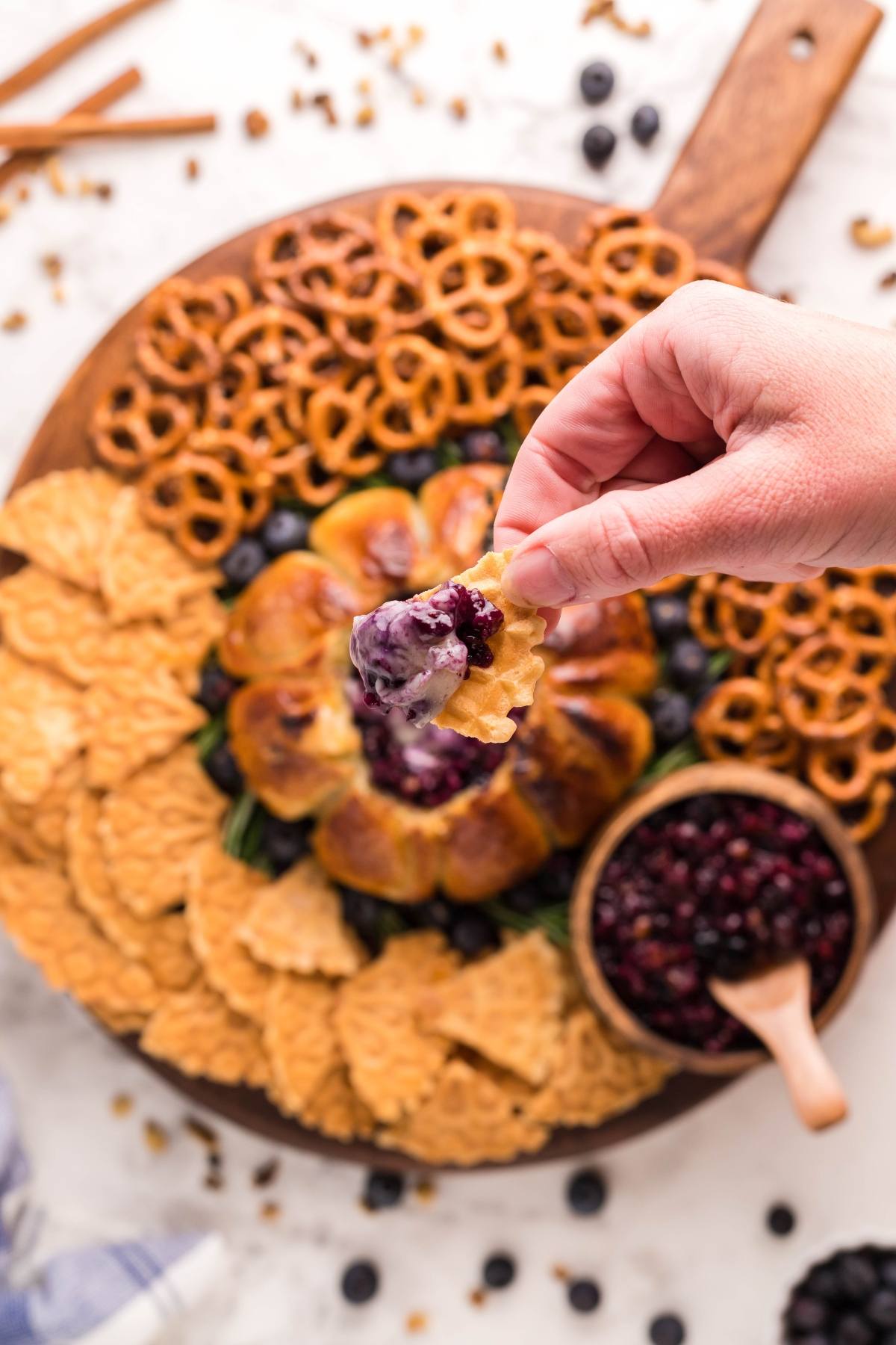 A hand dips a chip into blueberry brie appetizer dip on a platter with chips, pretzels, and fresh blueberries.