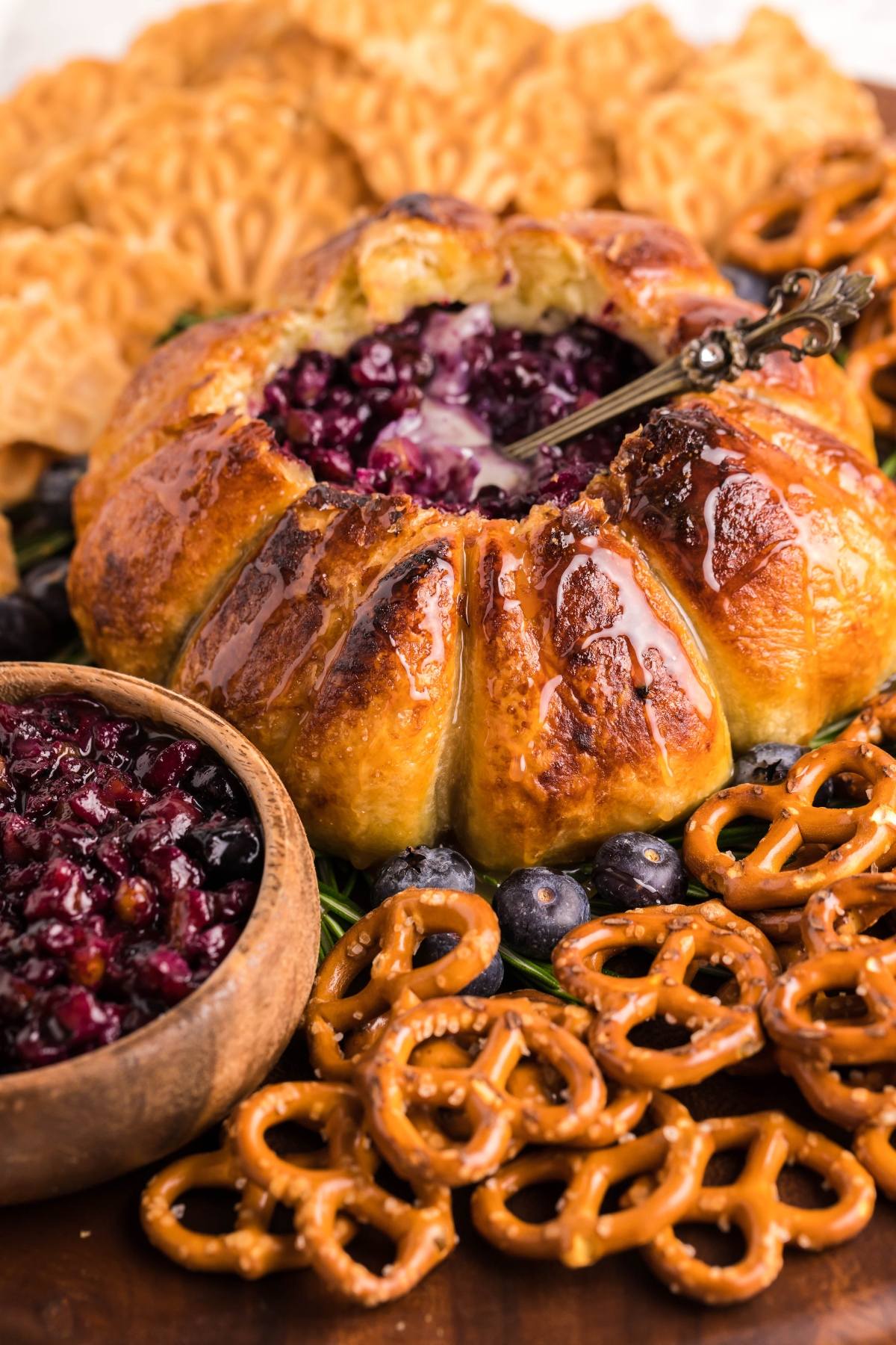 Air Fryer Baked Brie with Blueberries, featuring brie cheese dip with berry filling, is surrounded by pretzels, crackers, fresh blueberries, and a bowl of berry sauce for dipping.