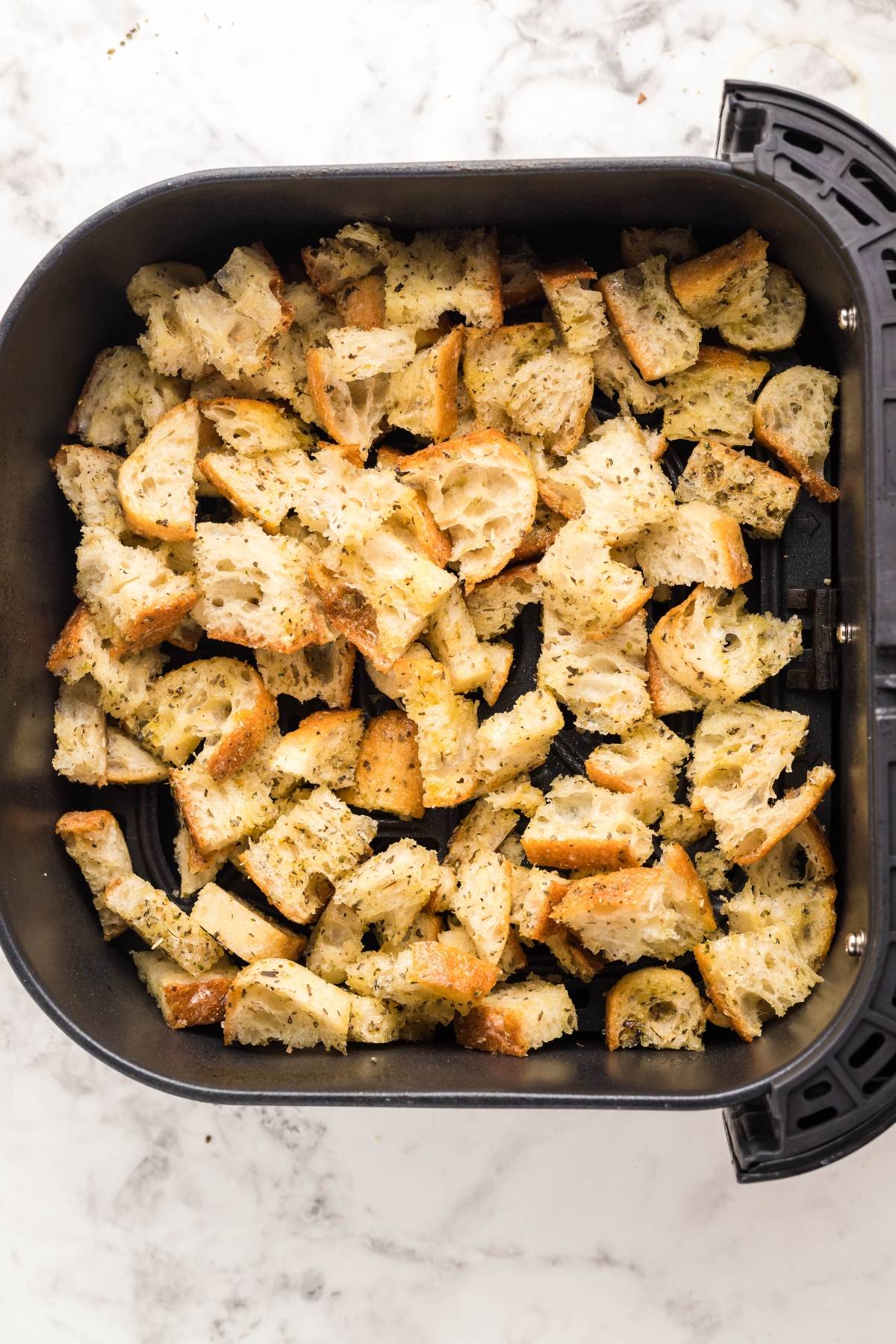 Cubed, seasoned bread pieces in an air fryer basket on a marble surface—perfect for making air fryer homemade croutons to top your salad with the best homemade Caesar dressing.
