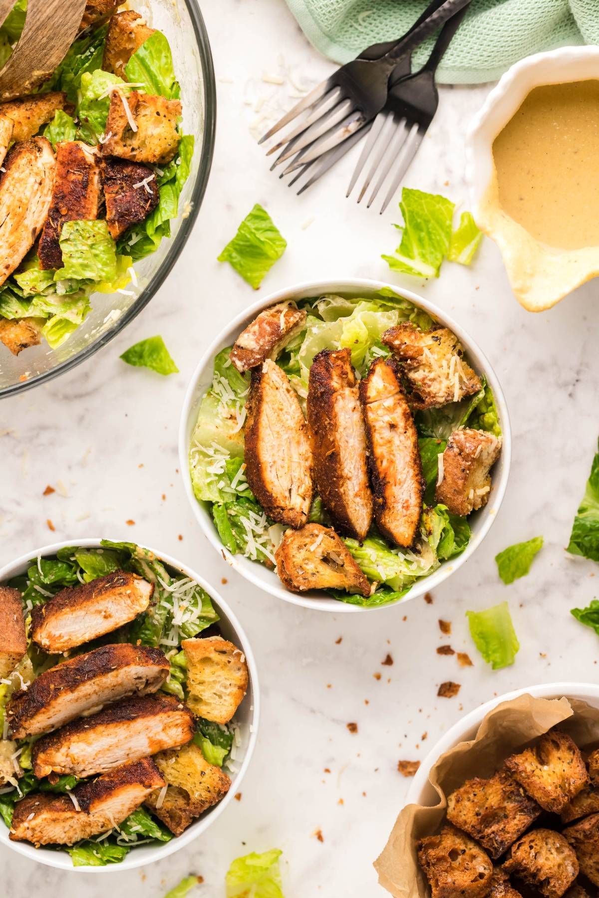 Bowls of Air Fryer Blackened Chicken Caesar Salad with air fryer homemade croutons, grilled chicken, and shredded cheese arranged on a marble surface.