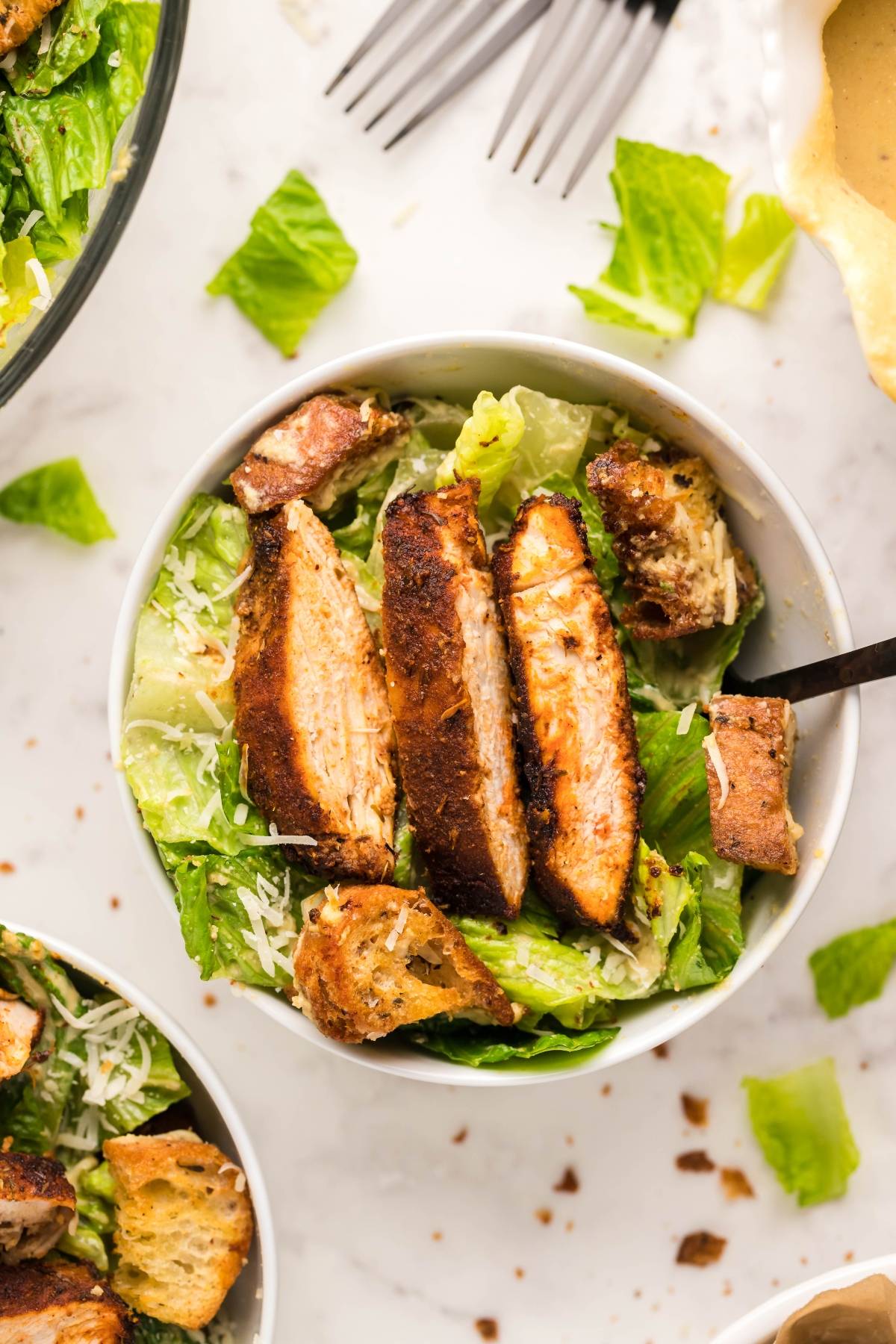 Bowl of Air Fryer Blackened Chicken Caesar Salad with juicy grilled chicken slices, air fryer homemade croutons, and grated cheese on a white surface.