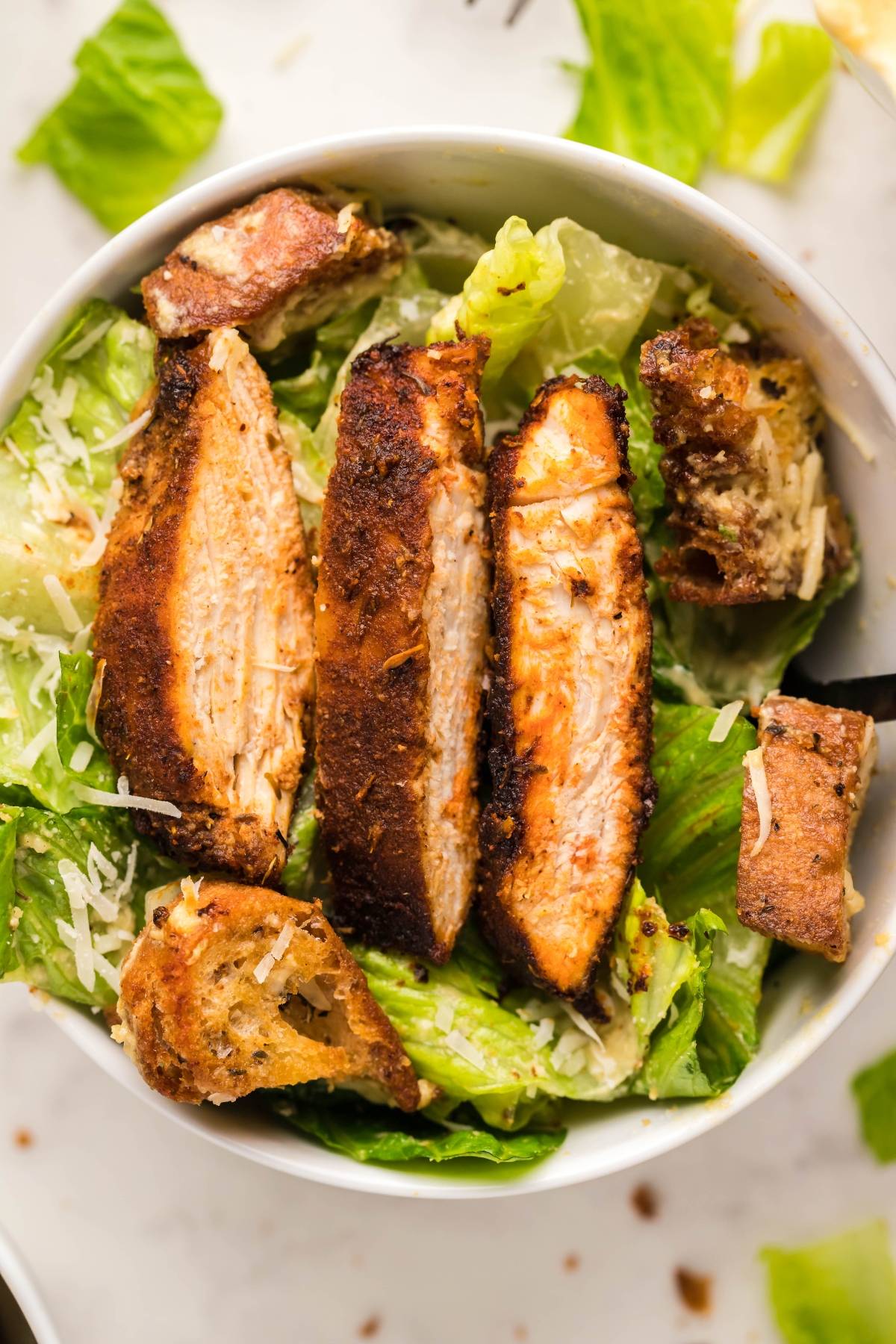 A bowl of Caesar salad topped with juicy grilled chicken slices, air fryer homemade croutons, and shredded parmesan cheese.