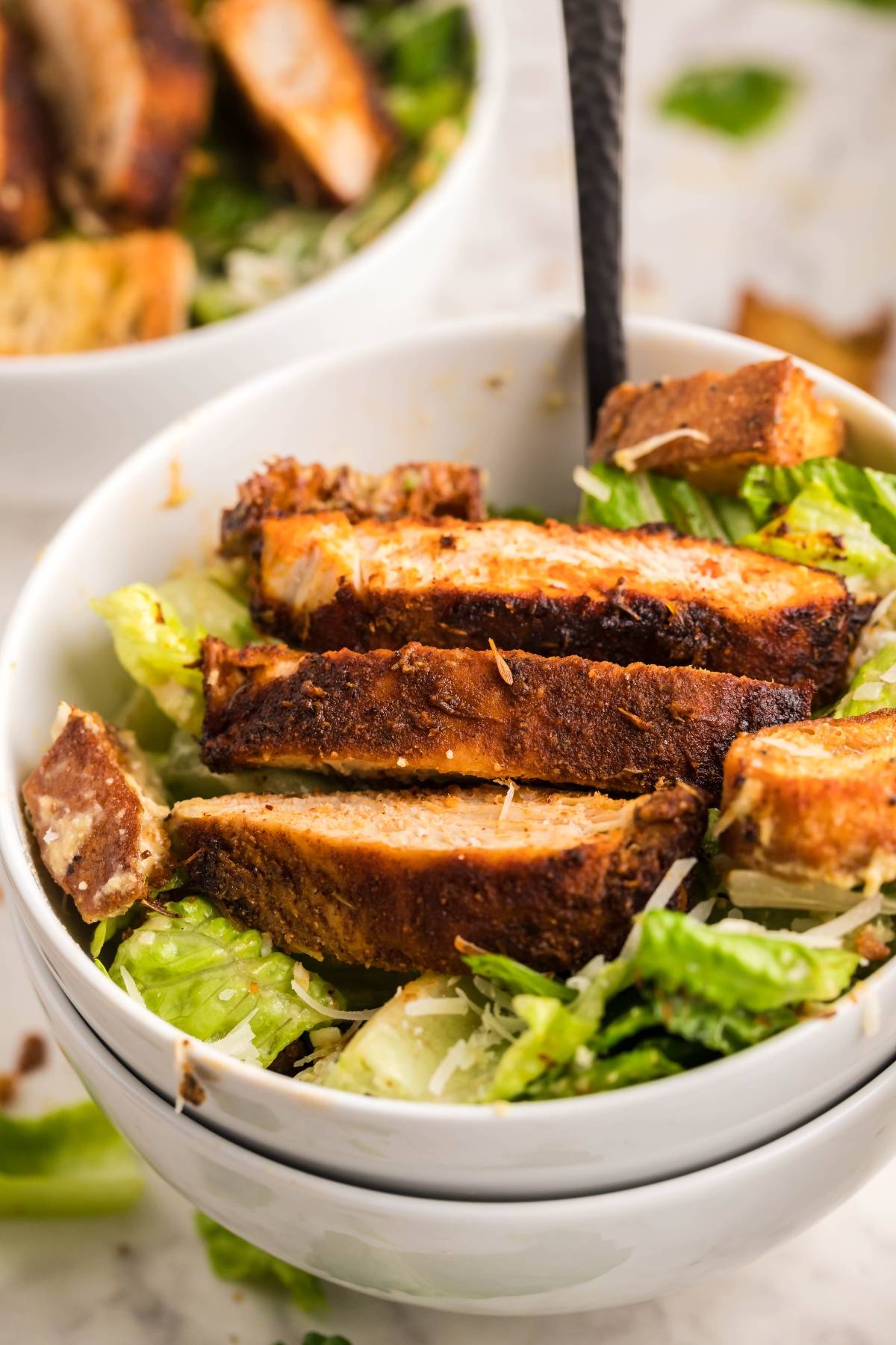 A bowl of Caesar salad topped with sliced, seasoned air fryer blackened chicken and croutons, all tossed with the best homemade Caesar dressing.