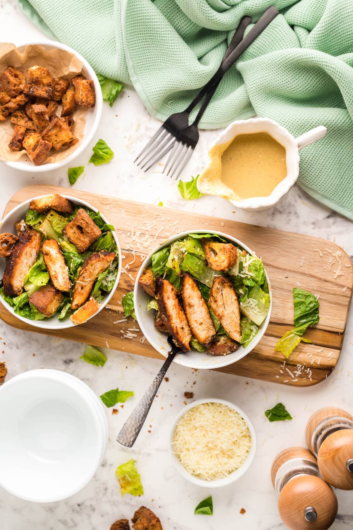 Two bowls of Air Fryer Blackened Chicken Caesar Salad on a board with croutons, the best homemade Caesar dressing, grated cheese, and utensils nearby—discover how to make blackened chicken in the air fryer for extra flavor.