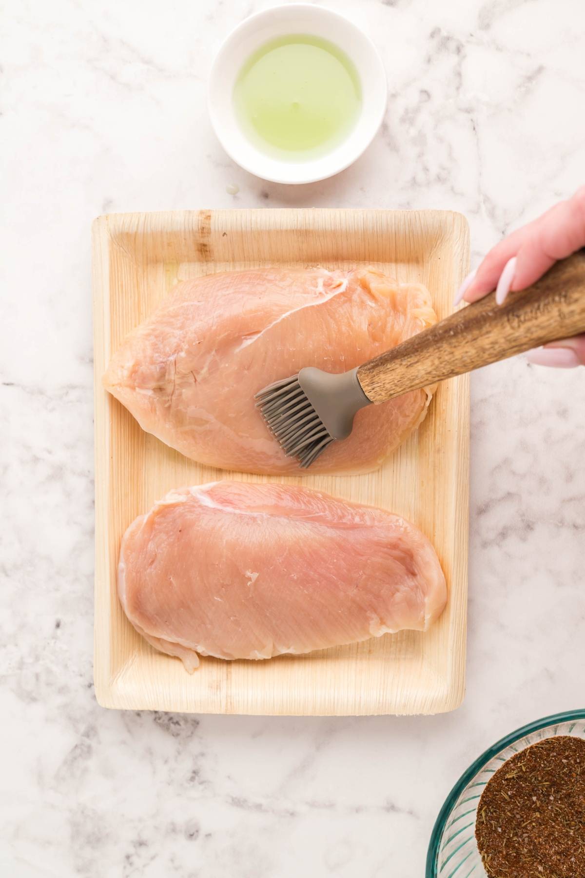 Two raw chicken breasts on a tray being brushed with oil, ready to become the star of an Air Fryer Blackened Chicken Caesar Salad, with a small bowl of oil nearby on a marble surface.