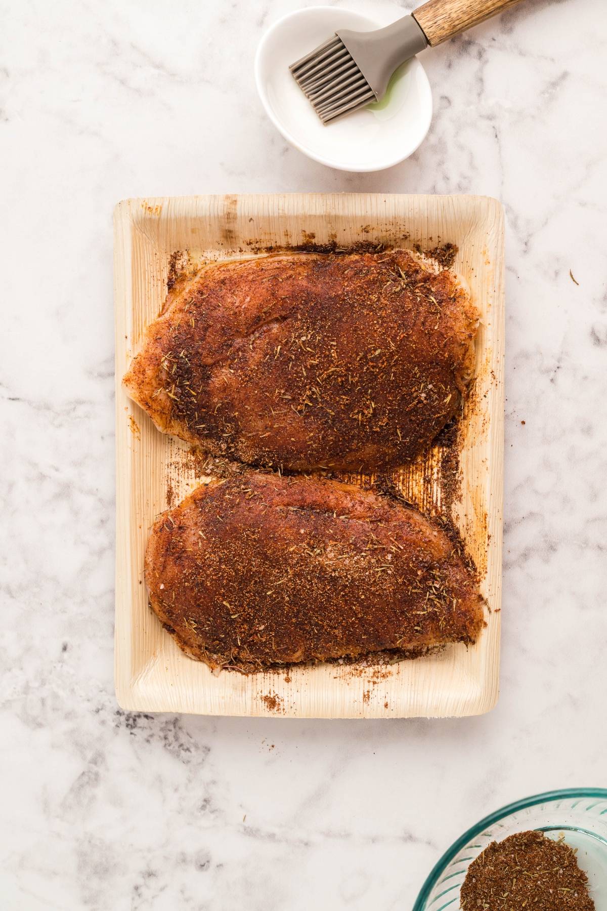 Two raw chicken breasts coated with spices on a tray, ready to become Air Fryer Blackened Chicken Caesar Salad. A basting brush and bowl are nearby—perfect for those learning how to make blackened chicken in the air fryer or the best homemade Caesar dressing.