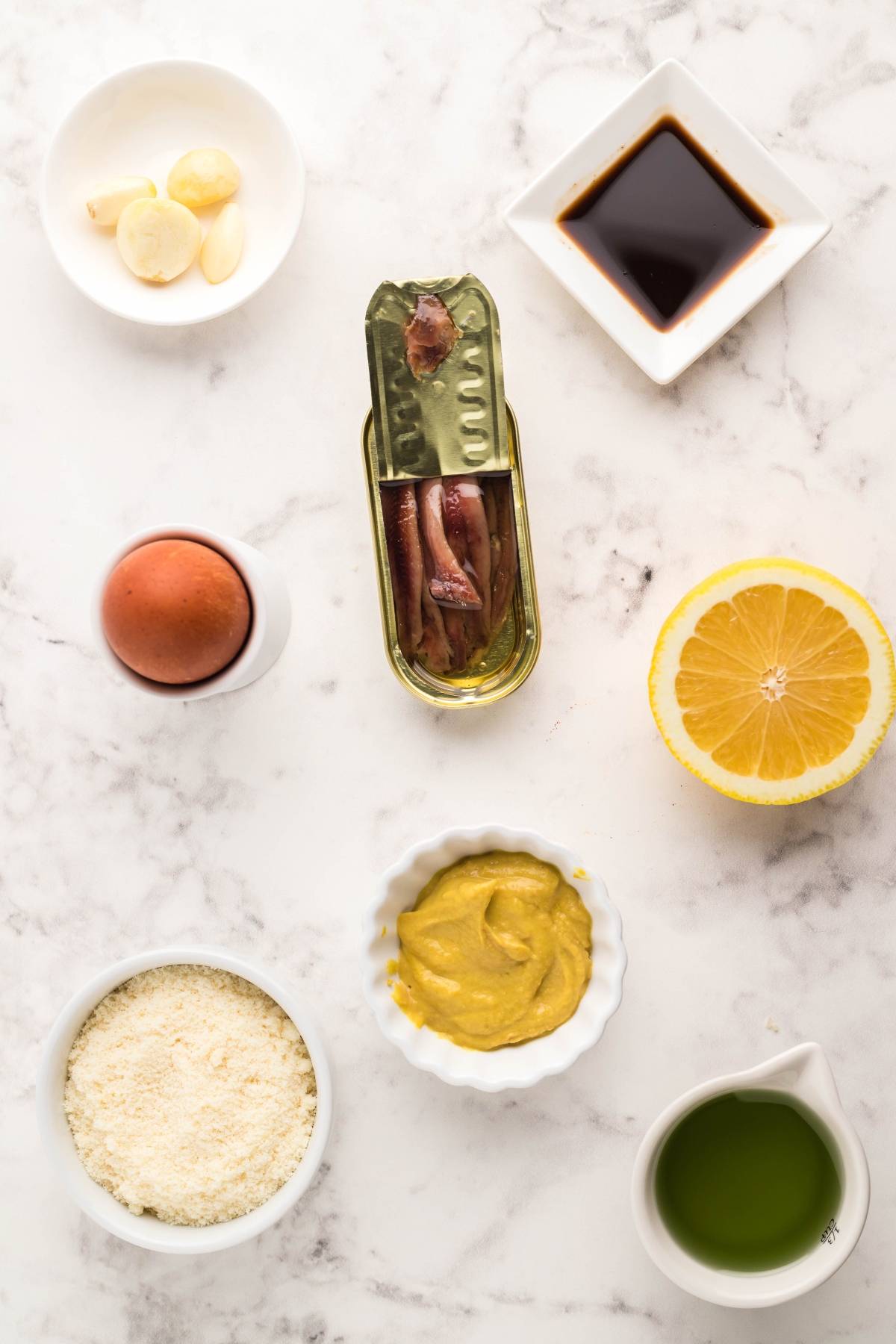 Ingredients for the best homemade Caesar dressing: garlic, soy sauce, egg, anchovies, lemon, Parmesan, mustard, and olive oil on a marble surface—perfect for an Air Fryer Blackened Chicken Caesar Salad.