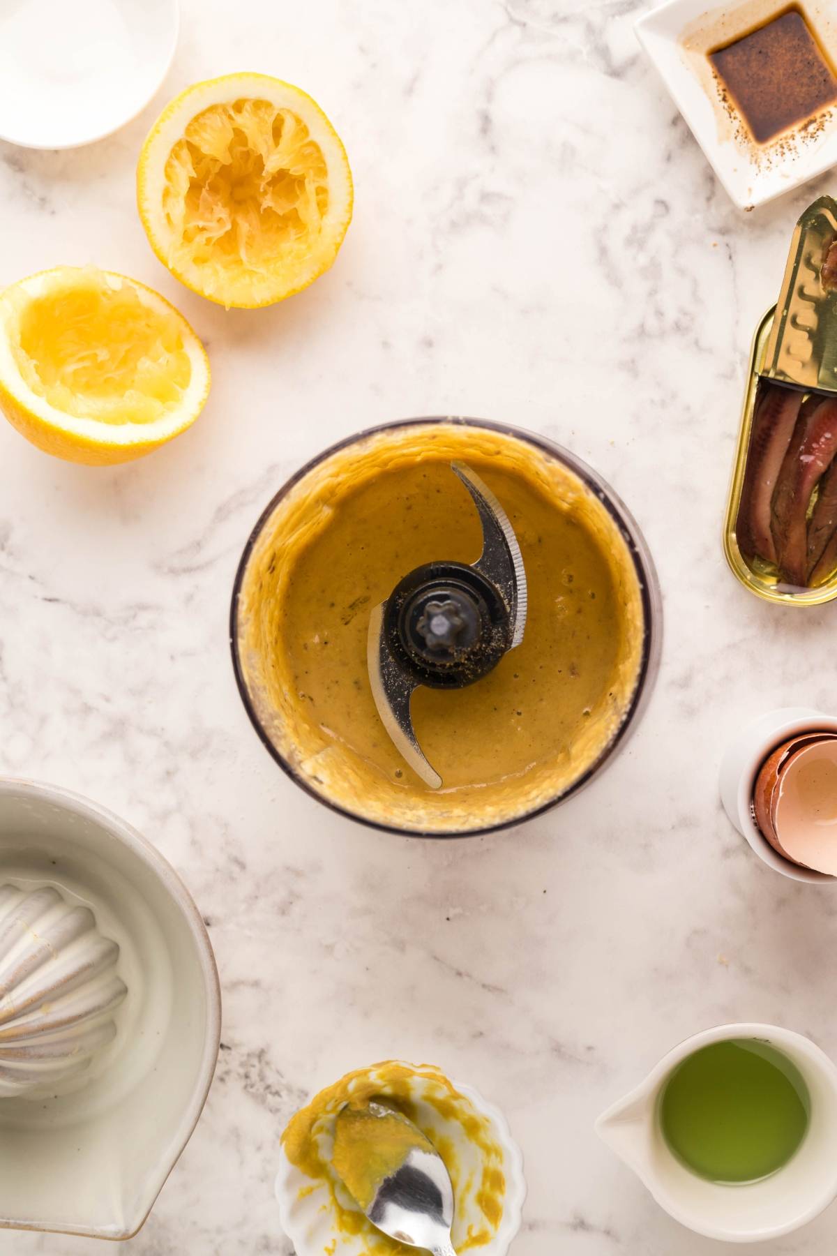 Food processor with creamy sauce, surrounded by lemon, anchovies, oil, and seasonings on a marble surface—perfect for topping Air Fryer Blackened Chicken Caesar Salad or tossing with air fryer homemade croutons.