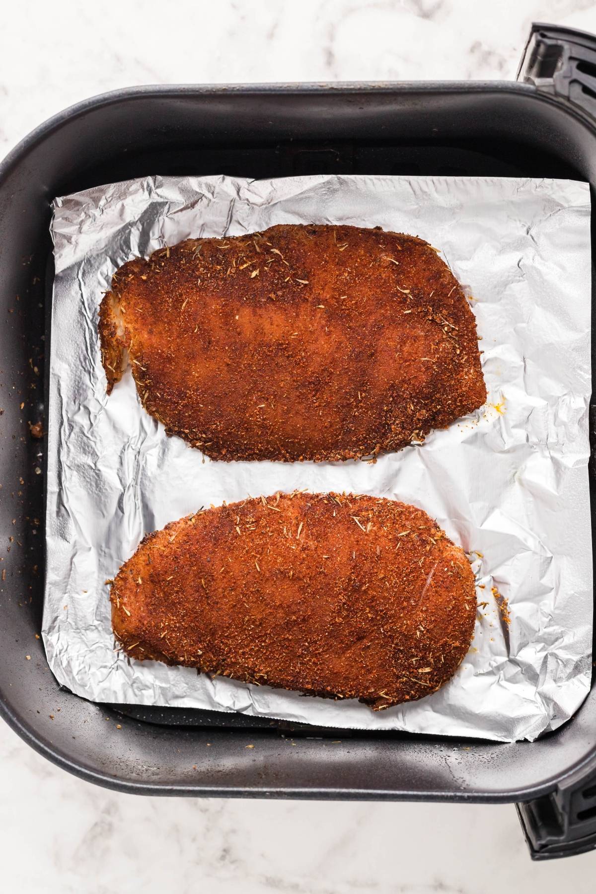Two breaded chicken breasts on aluminum foil in an air fryer basket, ready to be cooked—perfect for learning how to make blackened chicken in the air fryer or pairing with the best homemade Caesar dressing.