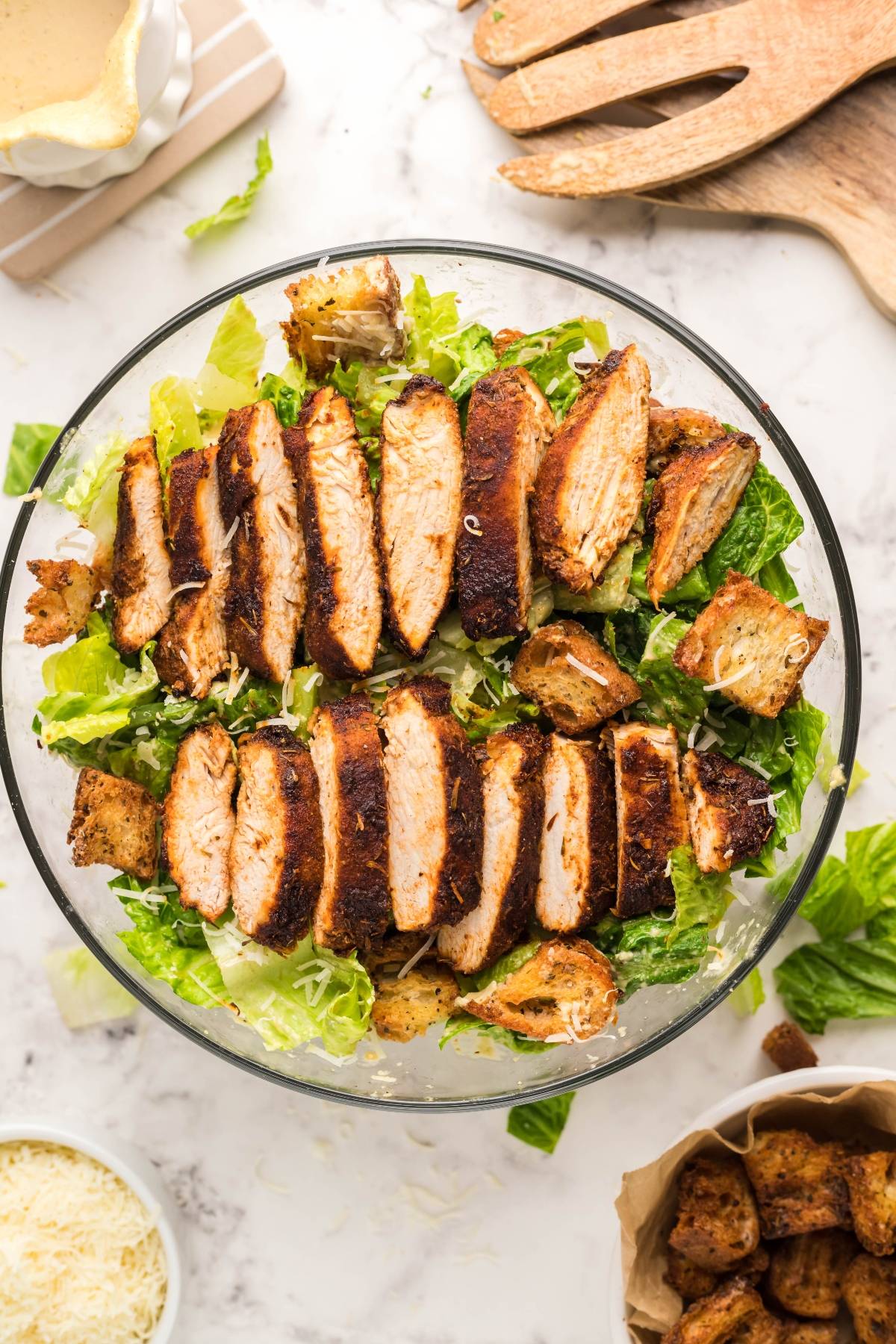 Sliced air fryer blackened chicken on romaine lettuce with air fryer homemade croutons and parmesan, topped with the best homemade Caesar dressing in a glass bowl.