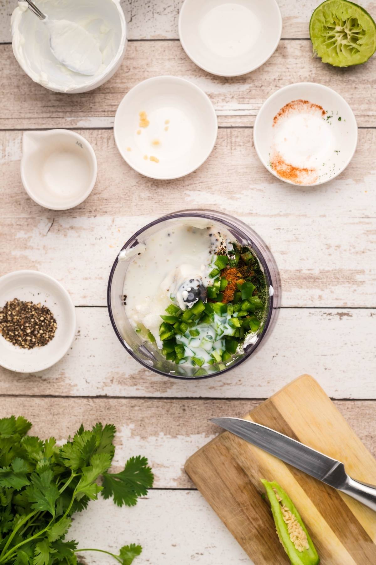 Top view of a food processor with herbs, yogurt, and spices—perfect for making Buttermilk firecracker ranch recipe—surrounded by bowls and fresh cilantro on a table.