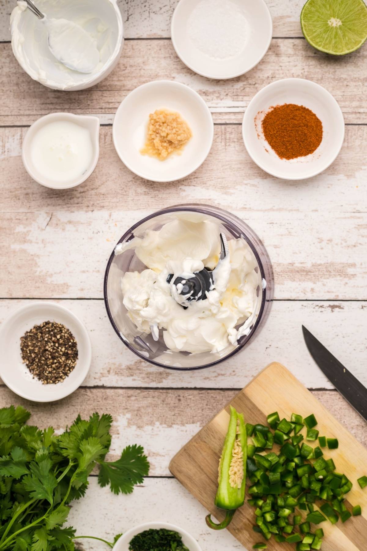 Overhead view of yogurt, spices, herbs, and chopped jalapeño on a table, with a food processor in the center—perfect for crafting a Homemade firecracker ranch dressing.