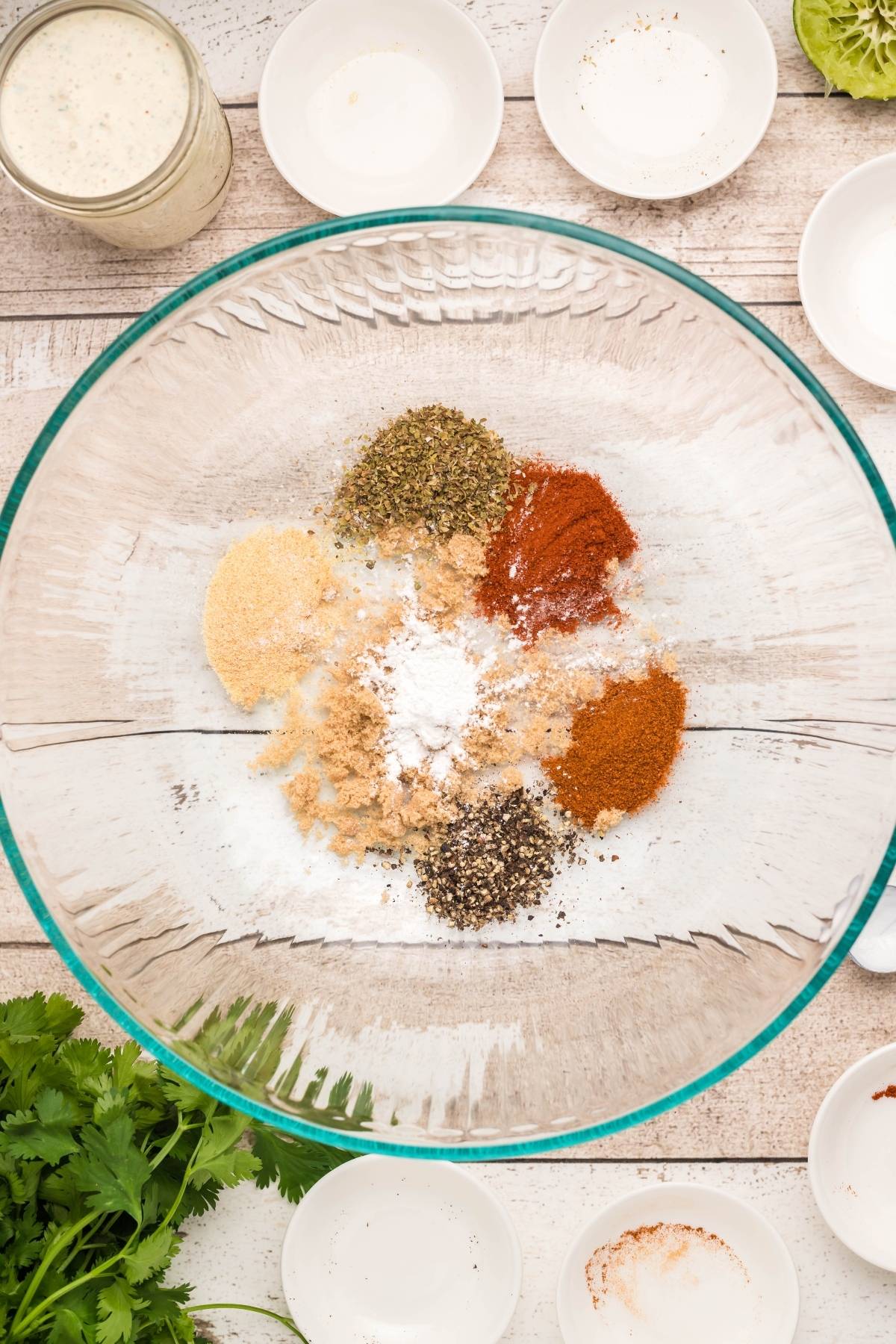 A glass bowl with various spices and seasonings—perfect for homemade firecracker ranch dressing—sits on a wooden table surrounded by empty small bowls.