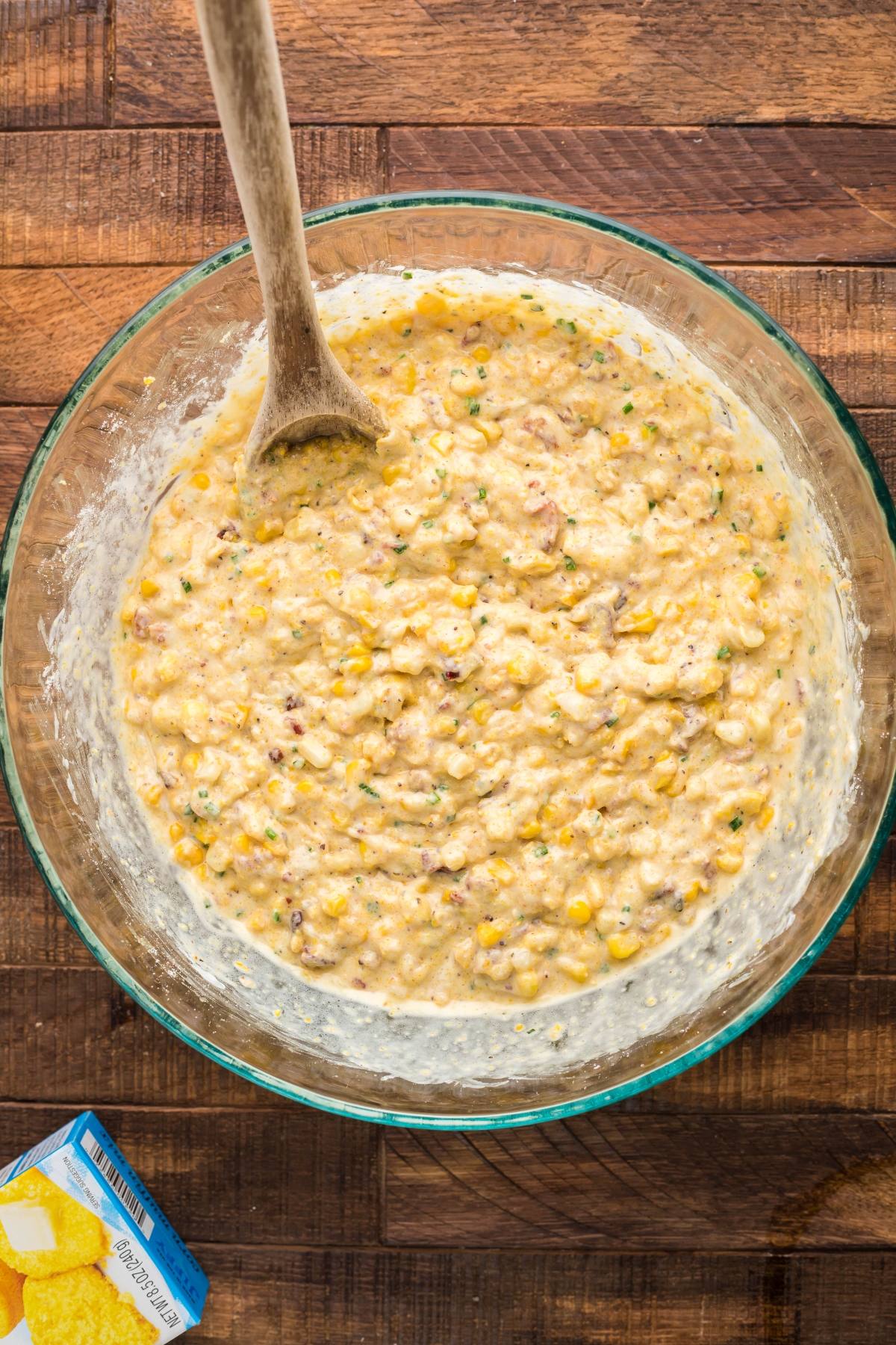 A glass bowl of corn fritter batter with corn, herbs, and bacon—perfect for a bacon corn casserole or even the best corn casserole made with Jiffy cornbread mix—stirred with a wooden spoon on a wooden table.
