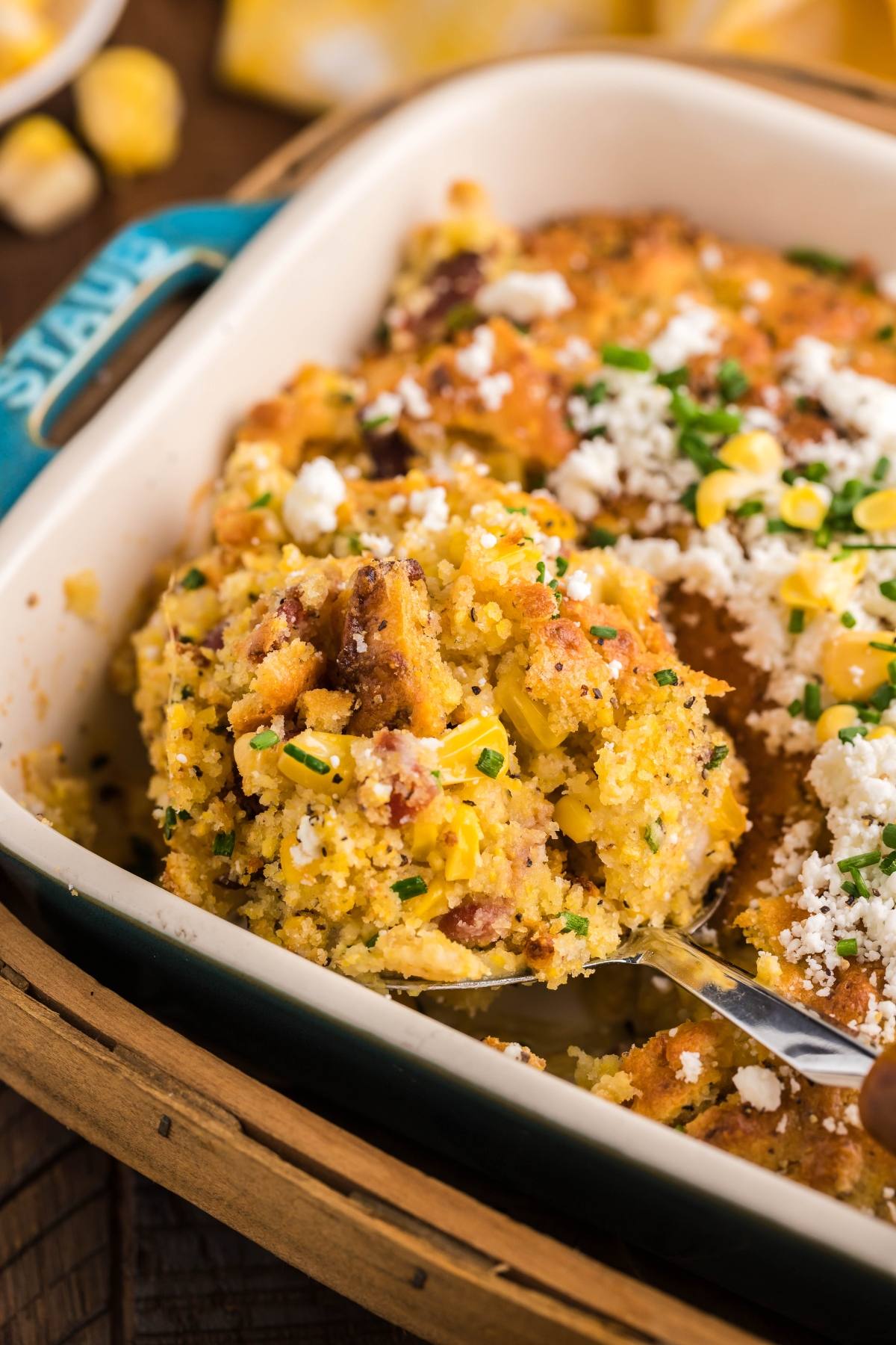 A spoon scoops cheesy bacon corn casserole with fresh herbs from a baking dish—perfect for those craving the best corn casserole.