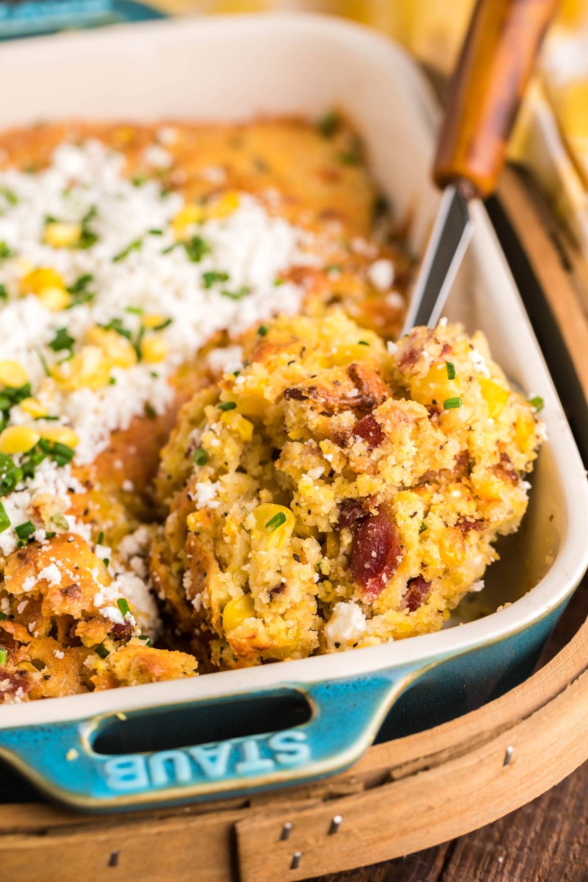 A serving of bacon corn casserole is being scooped from a blue Staub dish, featuring baked cornbread with bacon, corn, and cheese—perfect as an Air Fryer Corn Casserole twist on a classic favorite.