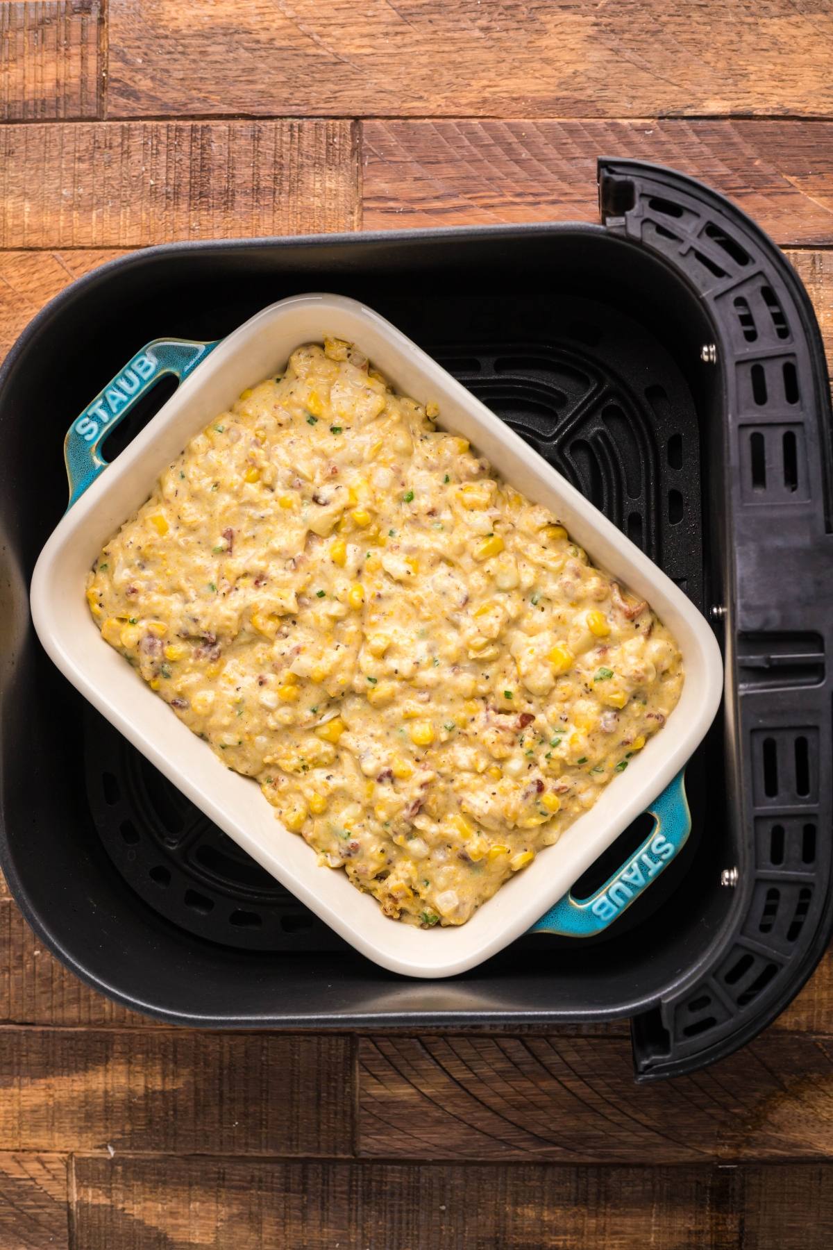 A casserole dish with Air fryer cornbread pudding made with Jiffy cornbread mix sits inside an air fryer basket on a wooden surface.