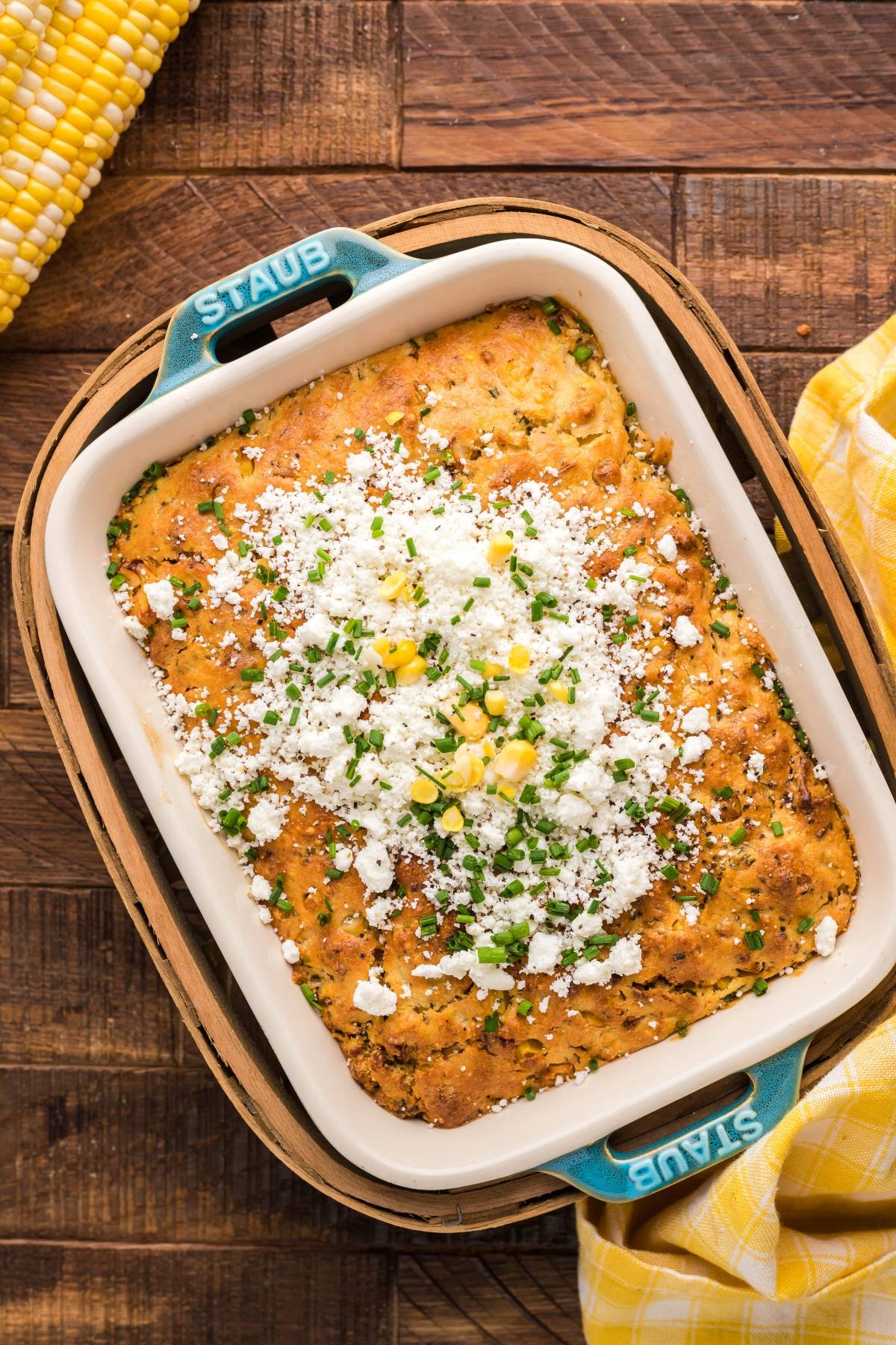 Baked cornbread in a dish topped with crumbled cheese, chives, and corn, made with Jiffy cornbread mix, on a wooden surface.