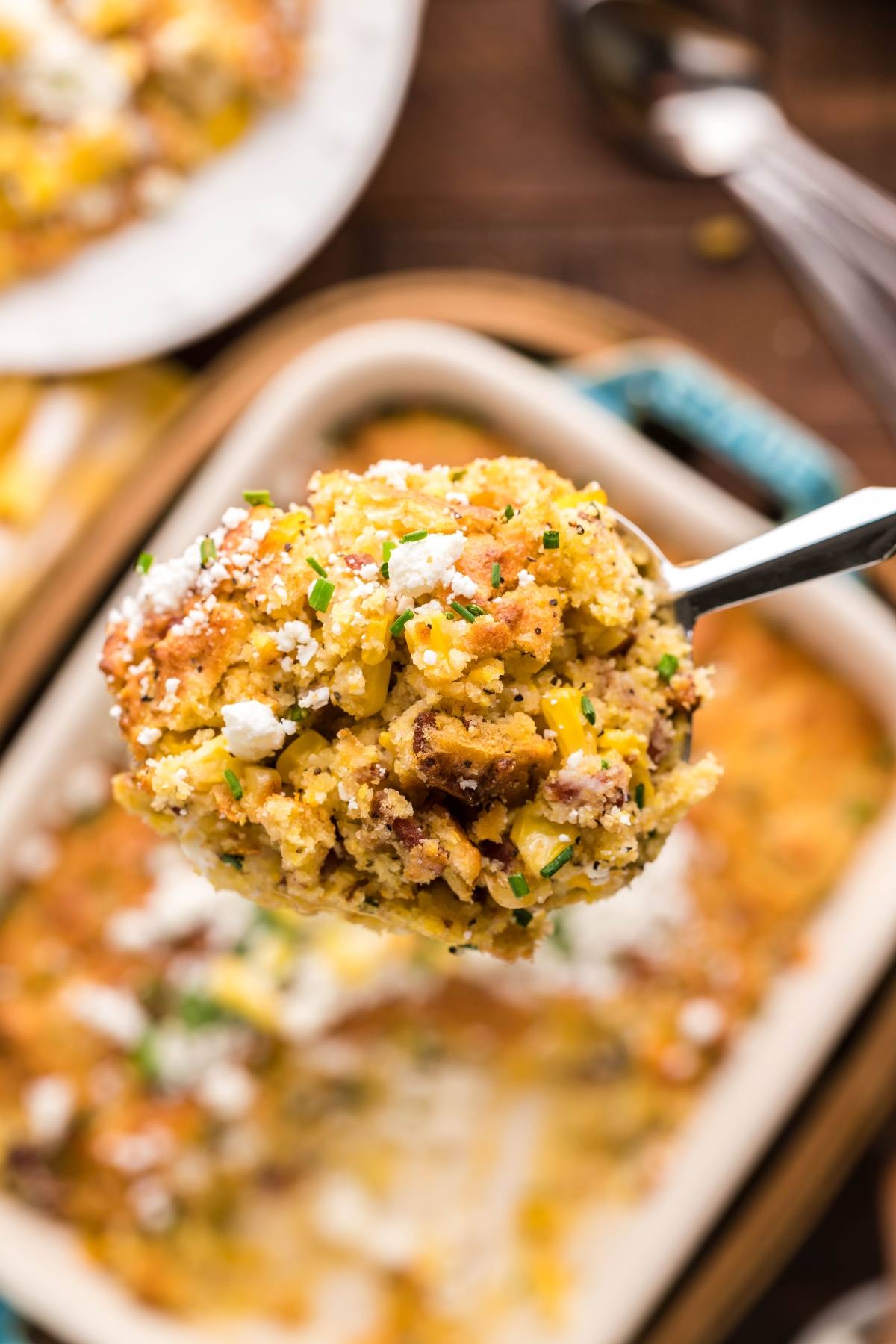 A spoonful of cheesy cornbread casserole, made with Jiffy cornbread mix, is held above a baking dish, sprinkled with herbs and cheese.