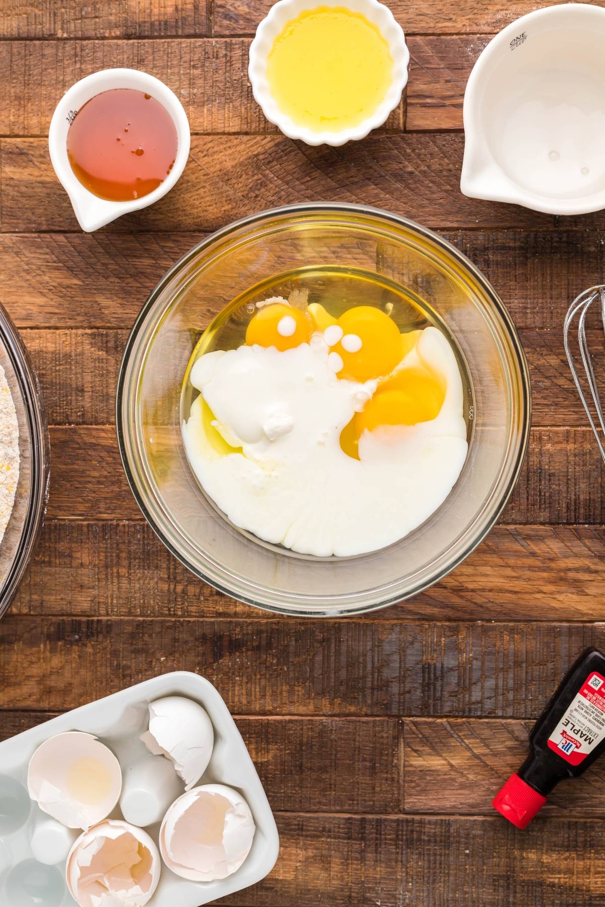 A bowl with eggs and milk, surrounded by vanilla, oil, cracked eggshells, and other baking ingredients—perfect for making Air Fryer Cornbread Cake with Honey Buttercream.
