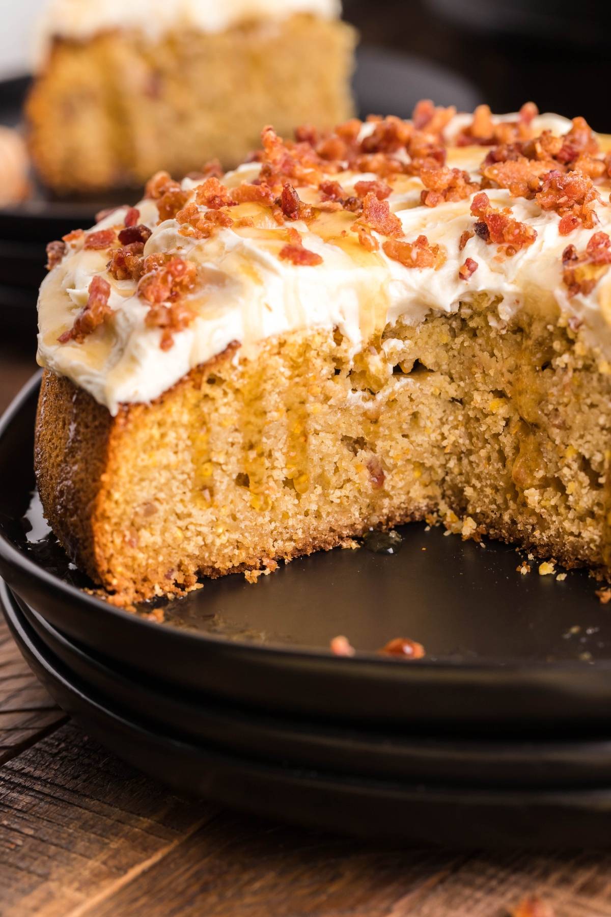 Close-up of an Air Fryer Cornbread Cake with Honey Buttercream frosting, topped with bacon bits, with a slice missing, on a black plate.
