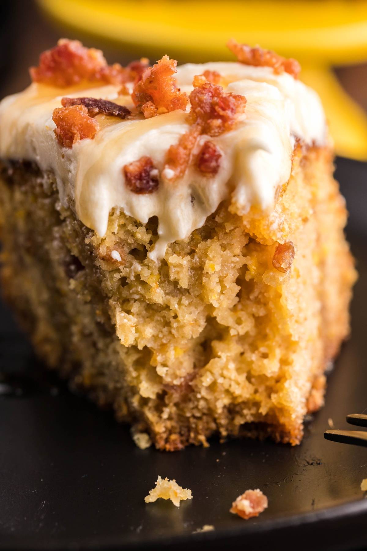 A slice of moist Air Fryer Cornbread Cake with Honey Buttercream and bacon crumbles sits on a black plate, offering the perfect balance of sweet cornbread with icing and savory flavor.