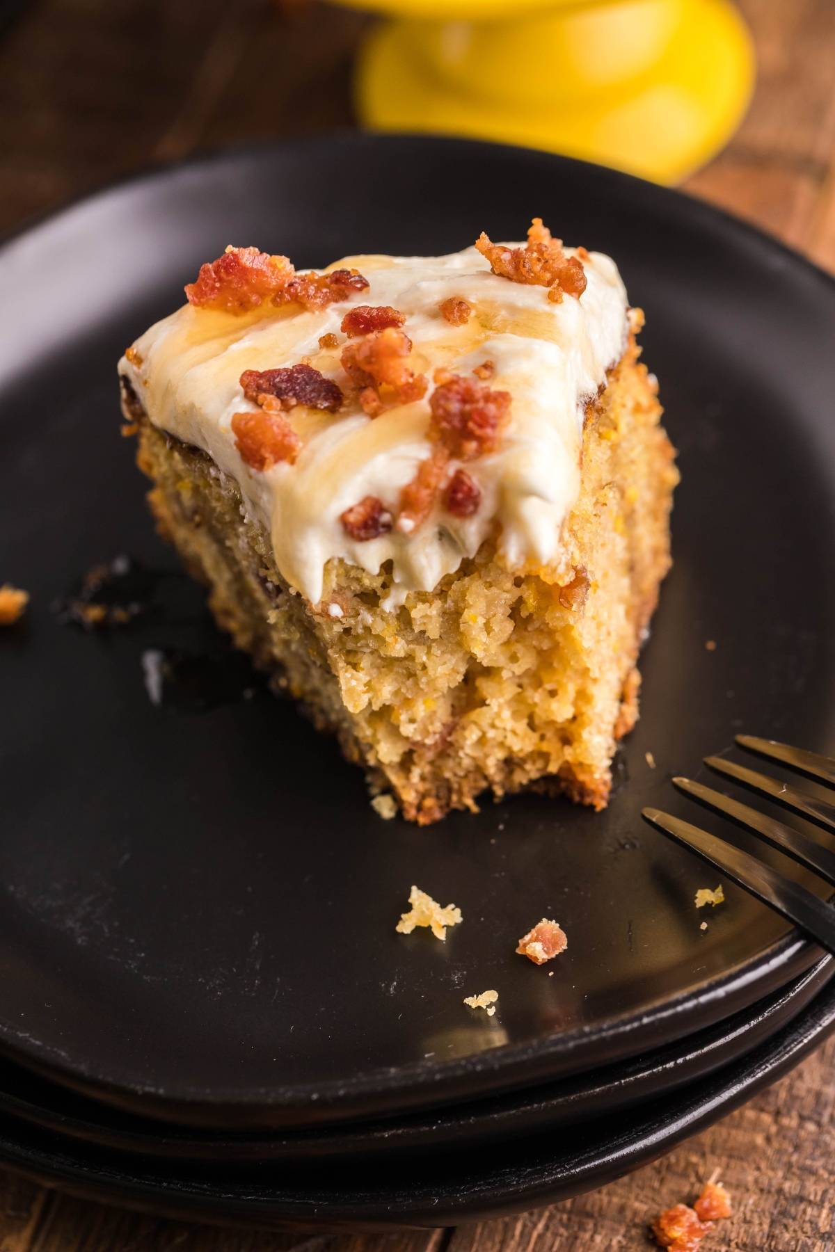 A slice of Air Fryer Cornbread Cake with Honey Buttercream frosting and crumbled bacon on a black plate, with a fork beside it.